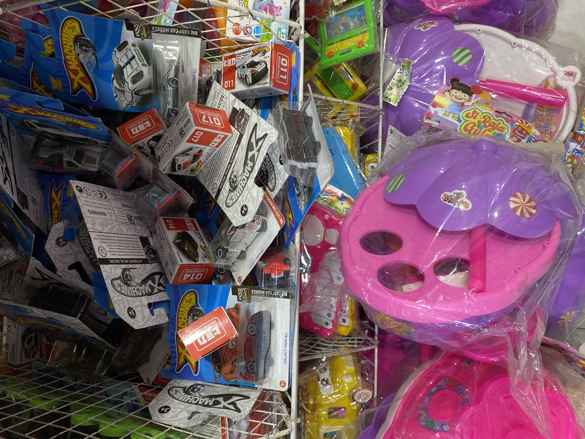 This image captures a close-up, eye-level view of a retail display at a store in Banda Aceh, Indonesia, likely a toy shop, filled with various toy cars and larger plastic toys. The primary subjects are toy vehicles, predominantly die-cast cars packaged in blister packs or small boxes, predominantly under the "X-Machines" brand. Some of these packages display graphics of racing cars, while others feature specific car models like a white and blue racing car. There are also red boxes with "TOMICA" branding visible. In the lower section of the image, larger plastic toys are displayed, including a bright yellow toy vehicle with cartoonish eyes, and two large, brightly colored plastic toys, one predominantly pink and the other purple, with visible plastic wrapping. The overall environment appears to be an indoor retail setting, with wire shelving units holding the merchandise. The lighting suggests an artificial light source, typical of a shop interior. No people are visible in the frame.