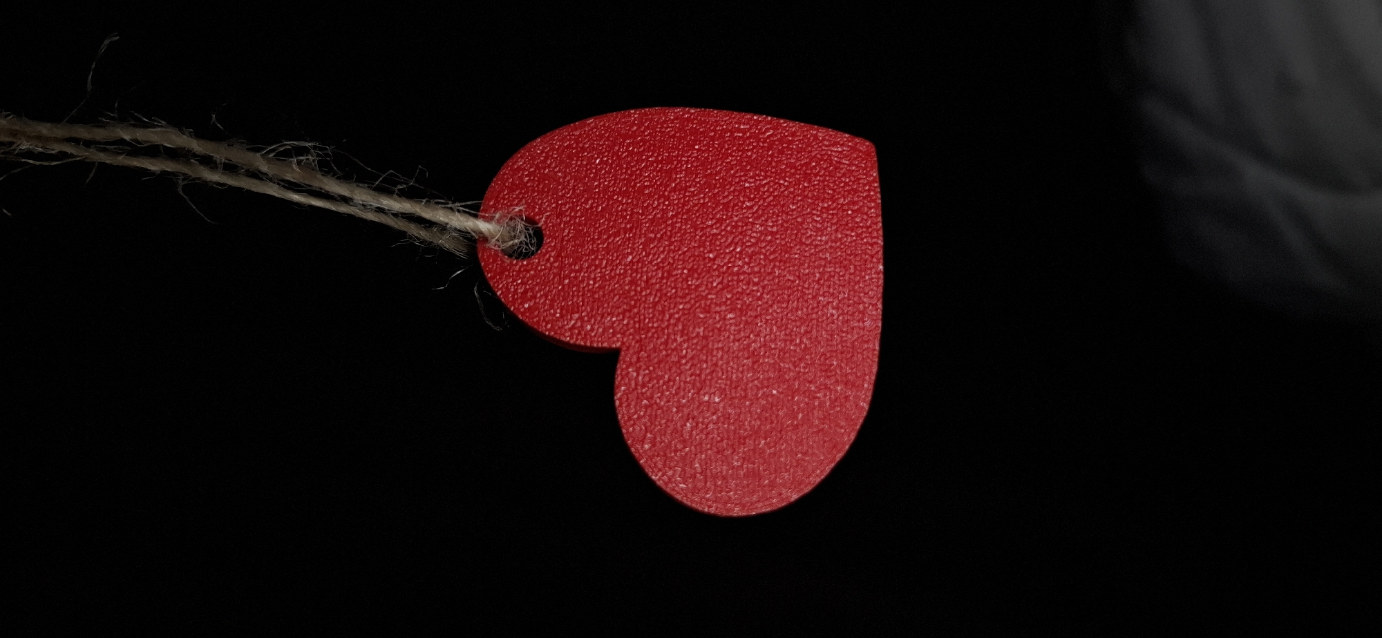 A single, red, heart-shaped object is suspended vertically against a plain black background. The heart possesses a textured, possibly granular or dimpled, surface. A small, circular hole is present near the top of the heart, through which a light-brown, multi-stranded piece of twine or natural fiber string is threaded. The twine shows visible individual fibers and slight fraying, extending upwards and slightly to the right from the heart's attachment point. The object is static, with no discernible actions or interactions occurring. No individuals are present in the scene. The image provides no visual information to determine its specific location within the city of Chaikovsky, Russia, or any other geographic context.