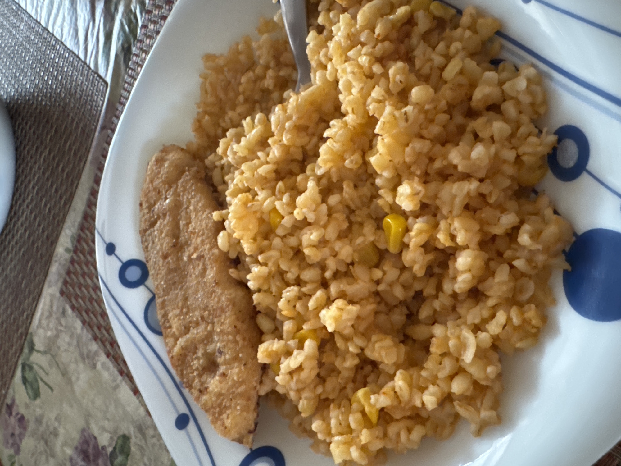 A plate of food, featuring a piece of breaded fish and a mound of rice with corn kernels, is presented on a patterned placemat. The plate has a white background with blue circular and linear designs. The scene is lit from above, casting subtle shadows on the food. The overall impression is of a meal prepared and served at a table, likely in a domestic setting.
