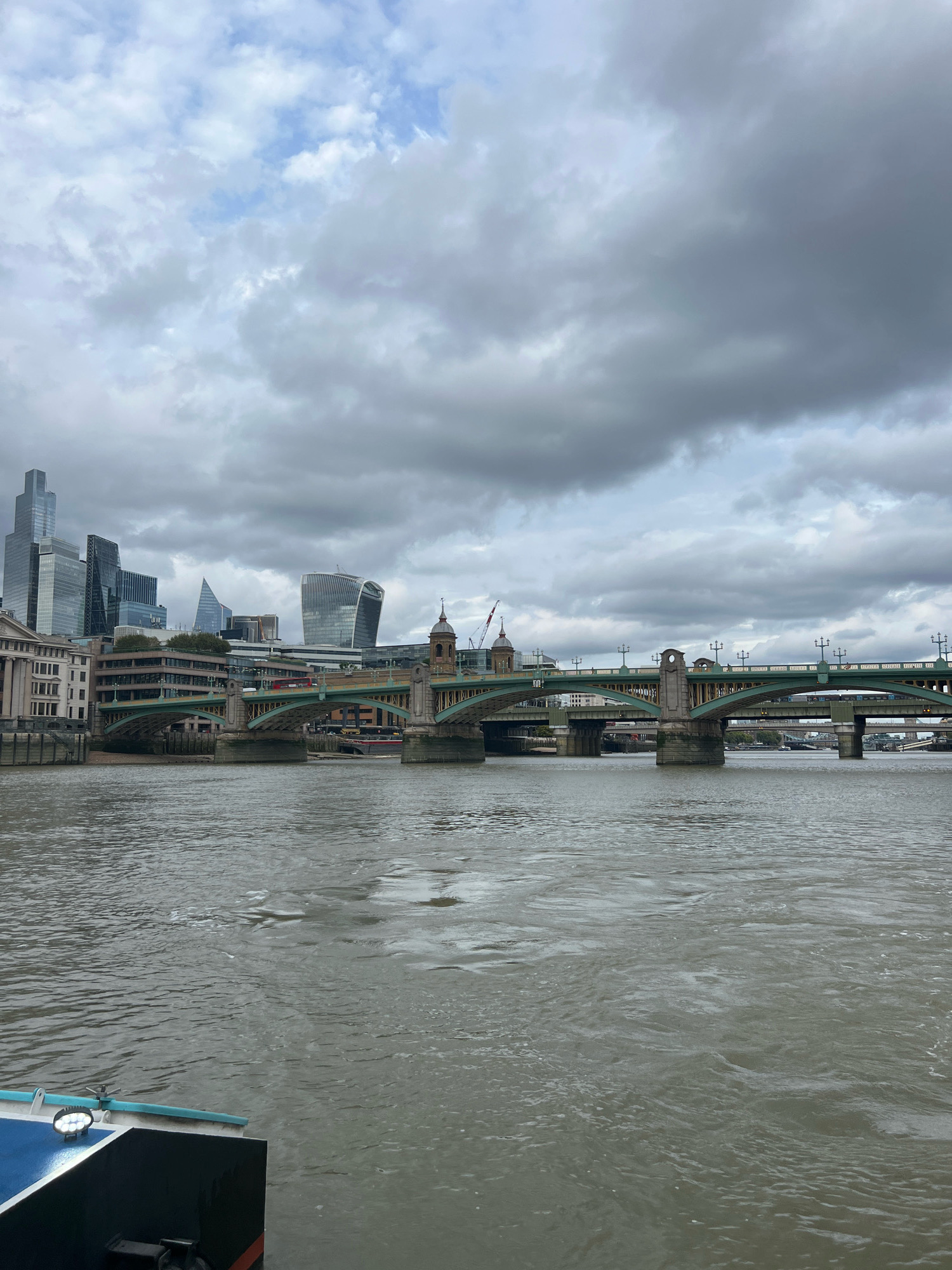 The image captures a view of the River Thames in London, United Kingdom, with the city skyline and a prominent bridge in the background. The sky is overcast with dramatic grey clouds, and the water of the river is choppy and a murky brown color.
In the foreground, the edge of a dark-colored boat with a blue and white section is visible on the lower left, featuring a spotlight.
The middle ground is dominated by a multi-arched bridge with green railings and stone piers. Beyond the bridge, the London skyline rises, featuring a collection of modern skyscrapers. Notable buildings include the Leadenhall Building (also known as the "Cheesegrater") with its distinctive angled shape, and the Gherkin building with its rounded form. Traditional stone buildings with neoclassical architectural elements are also present along the riverbank. The overall scene conveys a sense of urban grandeur under a somber sky.