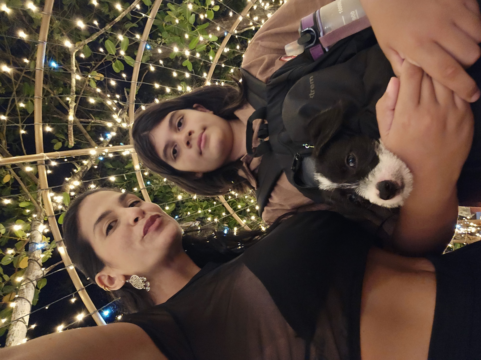 The image shows a woman and a child in front of a structure adorned with lights, possibly a pergola or trellis. The woman, who appears to be taking a selfie, is wearing a black top and has dangling earrings. The child is behind her, holding a black-and-white puppy. The location is likely Armação dos Búzios, Brazil, given the lights, foliage, and the camera angle used for selfies in this specific region.
