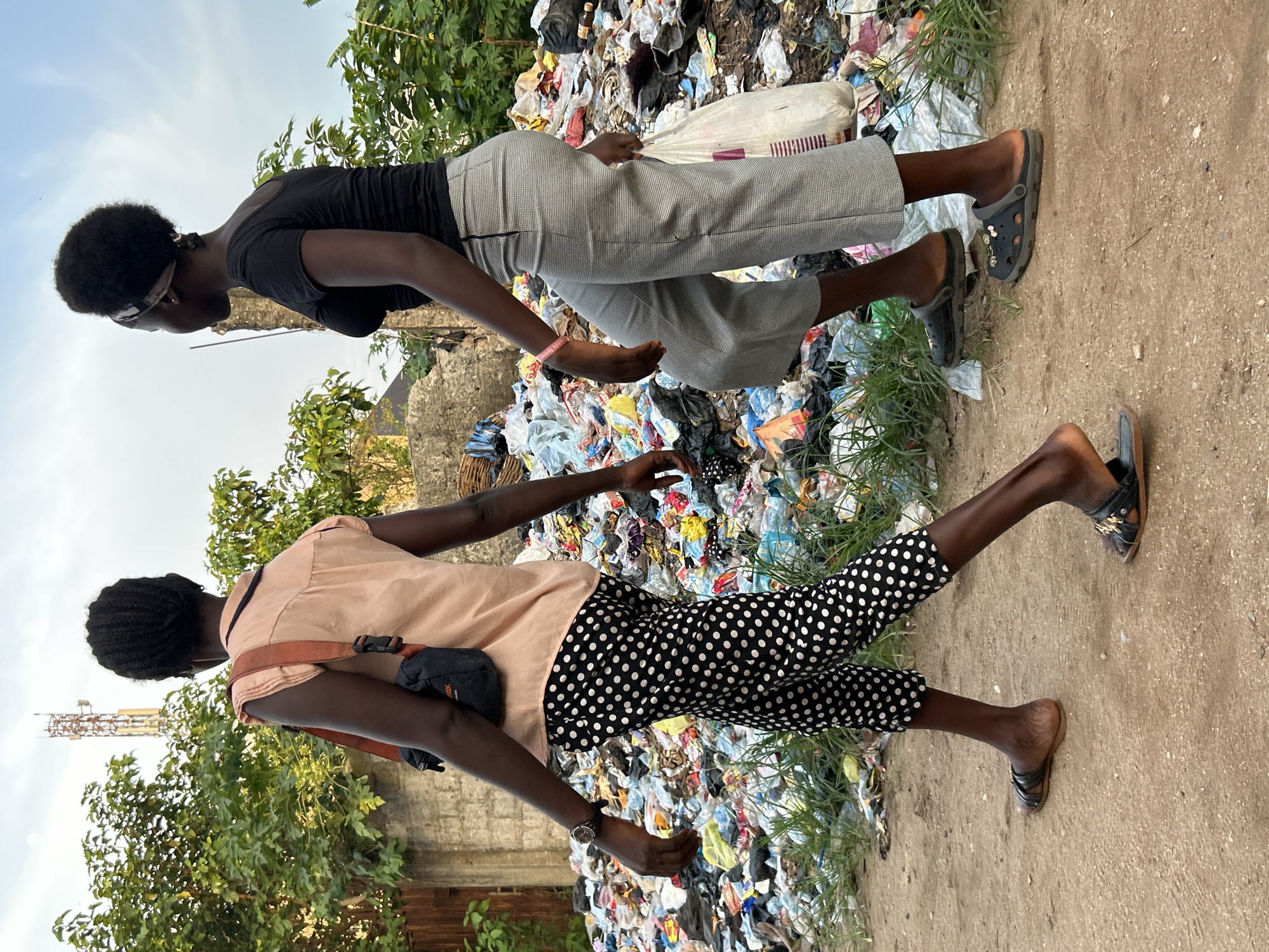 Two young adults walk past a large pile of garbage in Ihiagwa, Nigeria. The individual in the foreground is wearing a peach-colored short-sleeved shirt, black pants with white polka dots, and sandals. A black bag is slung across their shoulder. The individual in the background is wearing a black top, grey patterned pants, and black Croc-style shoes. The garbage pile is composed of mixed waste, including plastics and textiles. A dirt road borders the garbage pile. Vegetation is visible in the background. In the distance, a communication tower is seen. The sky is overcast.