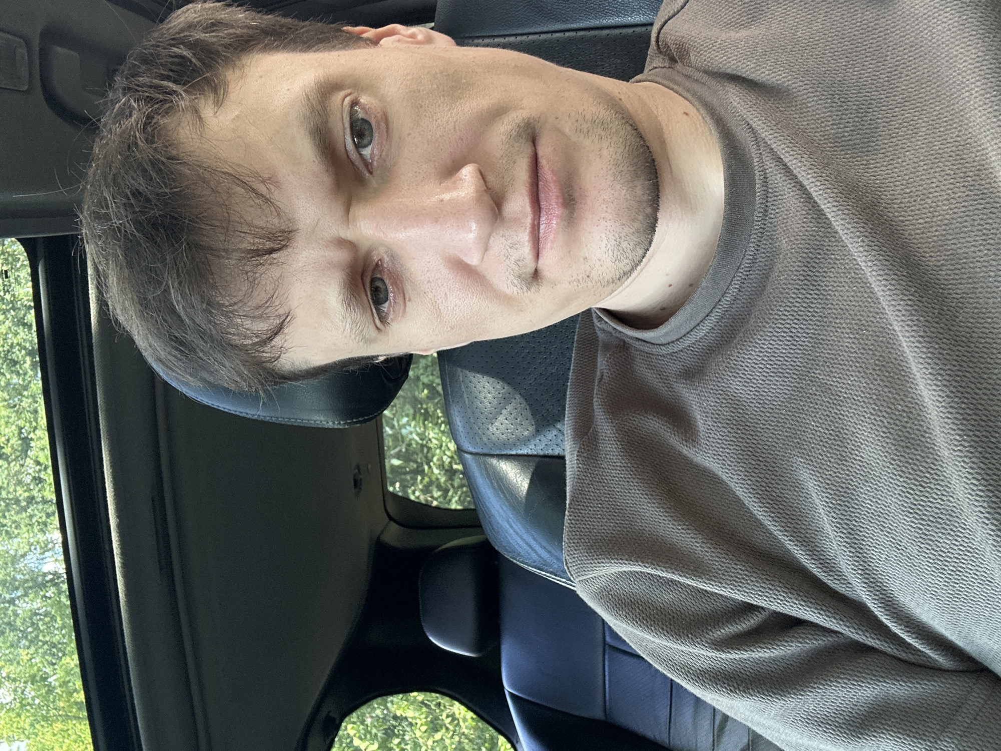 A selfie is taken from the chest up, showing a white man with short, dark brown hair and light brown eyes. He is wearing a brown, long-sleeved, textured shirt. His facial expression is neutral with a slight frown. The background is the interior of a car. The seats are black leather with perforated sections. A sunroof is visible at the top, with green foliage and sunlight filtering through. The man is seated in the driver's seat.