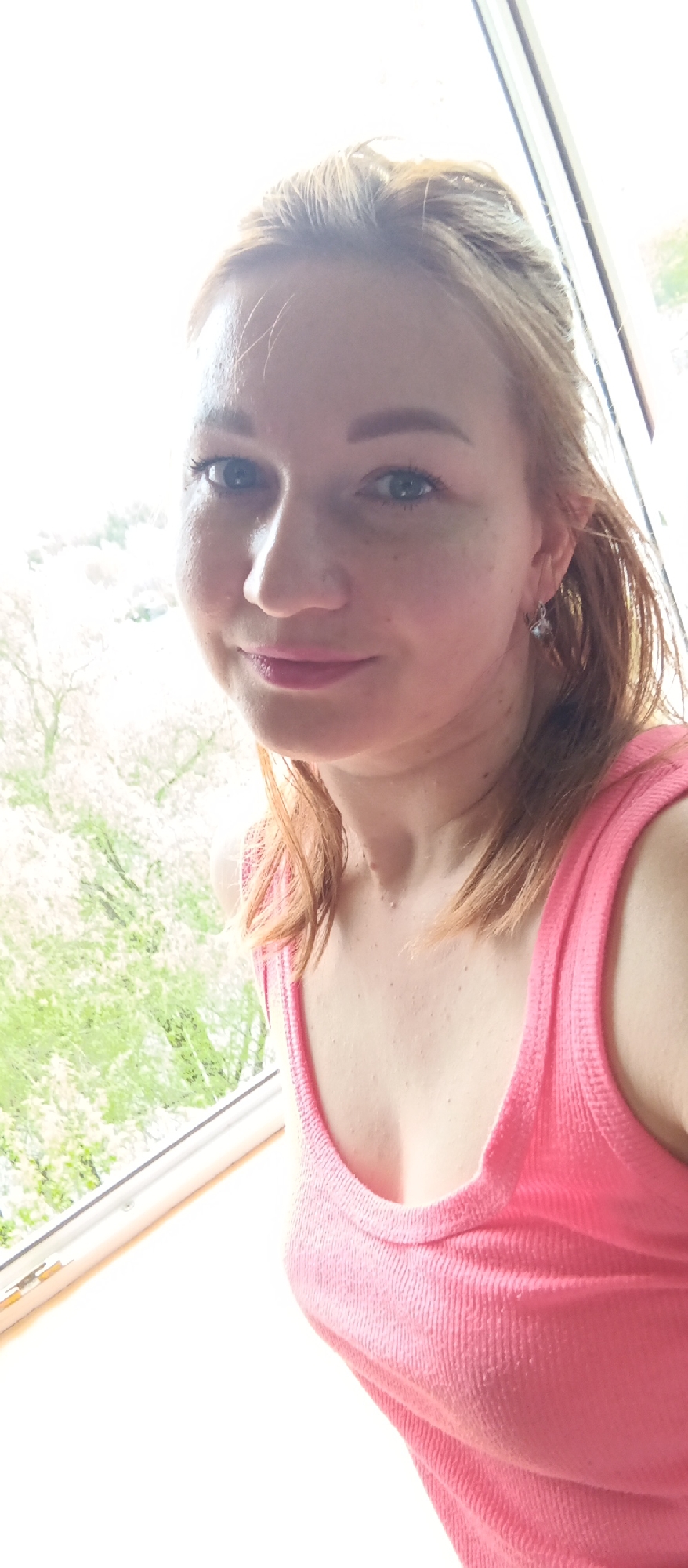 A woman is pictured indoors, likely near a window. She has fair skin, red hair, and blue eyes. She is wearing a pink tank top and earrings. Outside the window is a view of trees, possibly in bloom, with varying shades of green. The location is Unknown, Unknown.