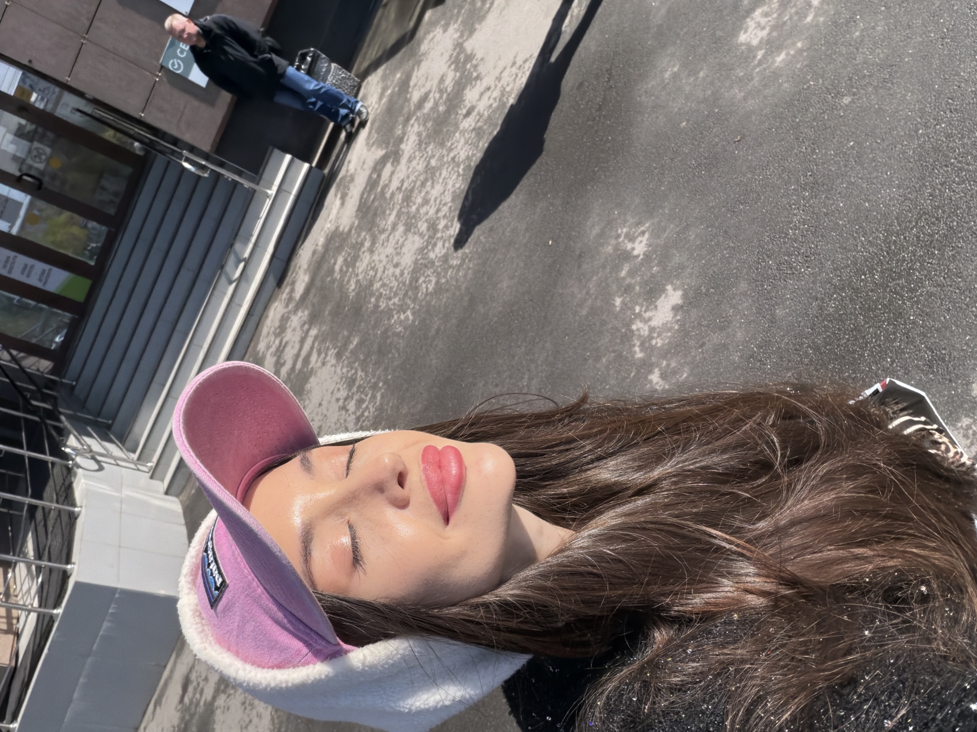A young woman lies prone on a gray asphalt surface, her face upturned toward the sun. She is wearing a pink baseball cap with white faux fur trim. Her eyes are closed, and she has lipstick applied in a heart shape. Long, dark hair fans out around her head. A shadow stretches across the pavement behind her, and the top of a building is visible on the left, with a man standing in front of it in the background. Based on the building style, the scene appears to be in Saint Petersburg, Russia.