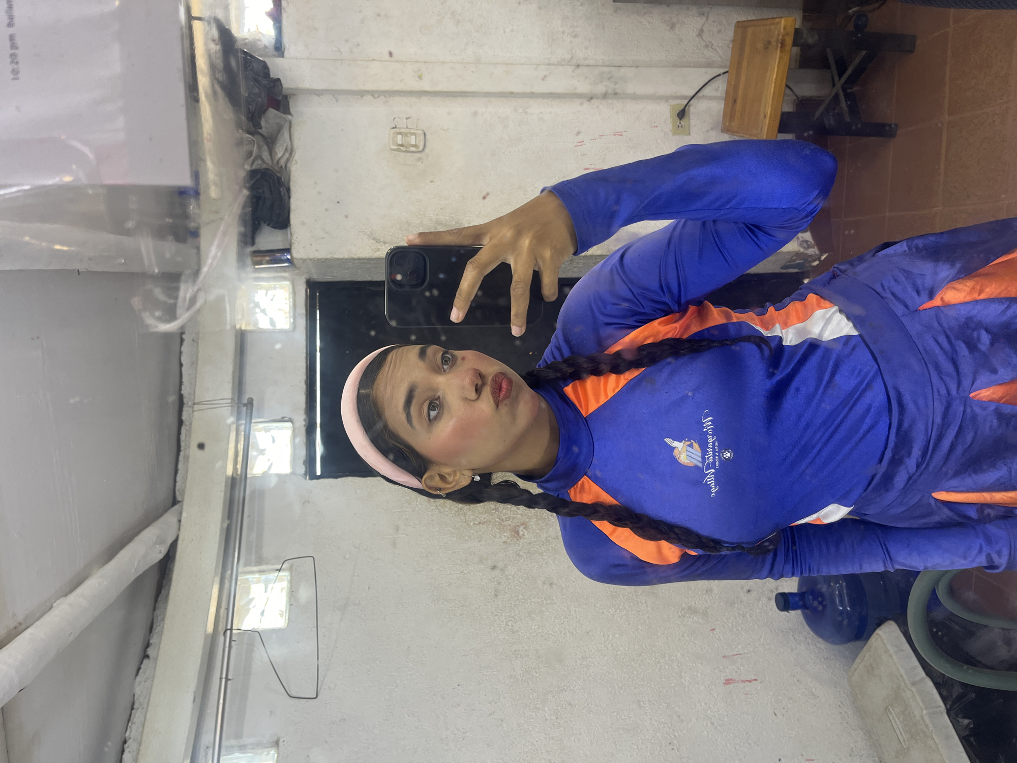 The image depicts a person in blue and orange athletic attire, possibly a uniform, with their dark hair in braids and wearing a pink headband. They are holding a smartphone, seemingly taking a selfie, with a slightly pursed expression. The setting appears to be an interior space with aged, off-white walls. The logo on their attire reads "Multiverse of Worlds" with a graphical symbol above it. In the background, a wooden stool, a glimpse of a possible water bottle, and some clear acrylic displays are visible. Given the "Multiverse of Worlds" logo and the context of Porlamar, Venezuela, this may be related to athletic or cultural events that are supported through that company in Porlamar.