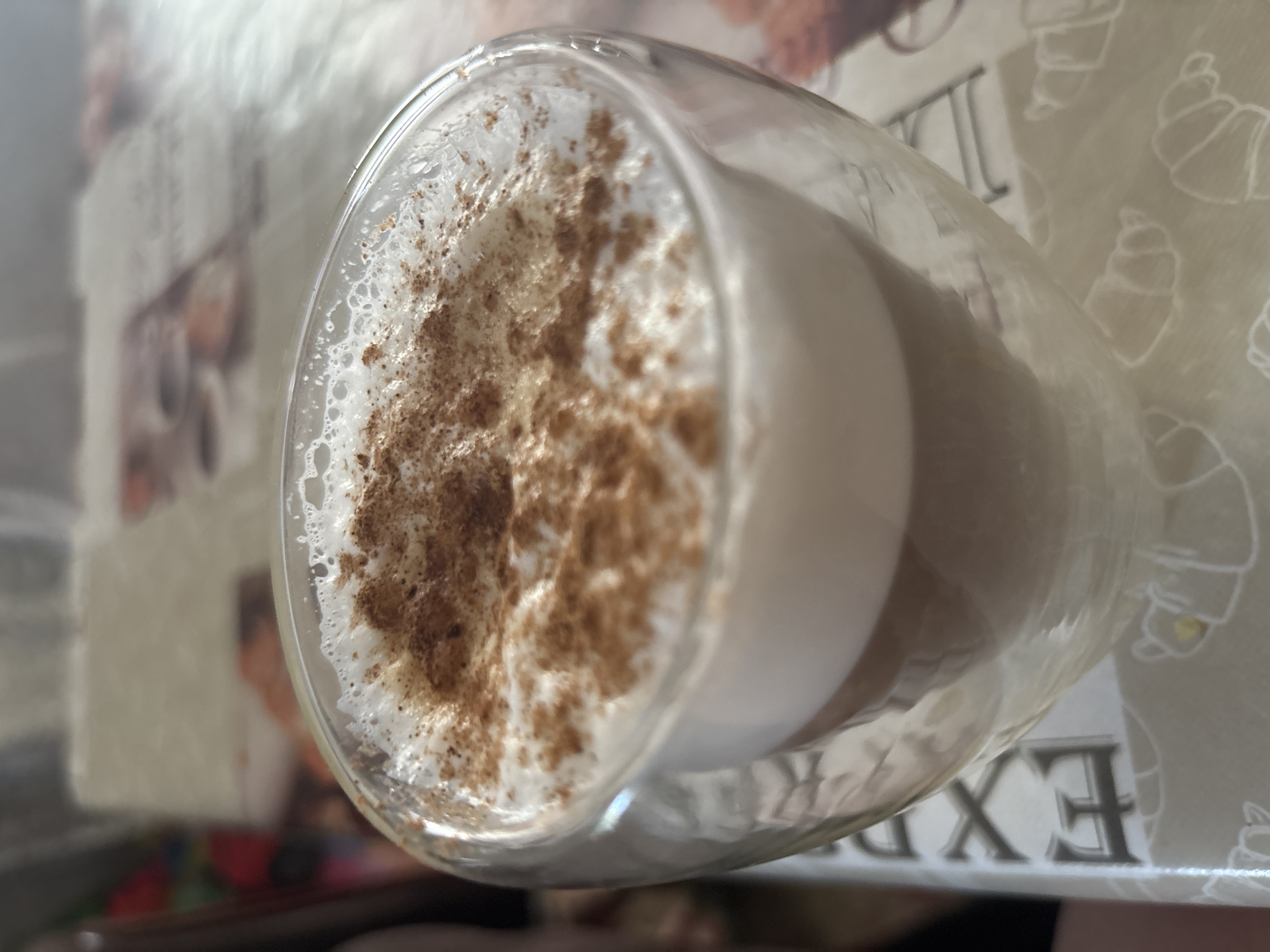 A double-walled glass mug filled with a latte and topped with frothed milk and cinnamon powder sits on a table. The mug is clear glass with a visible layer of the latte beneath the foam. The table surface has a patterned tablecloth with images of croissants. The background is blurred, showing hints of a room and possibly another person. The scene is indoors.