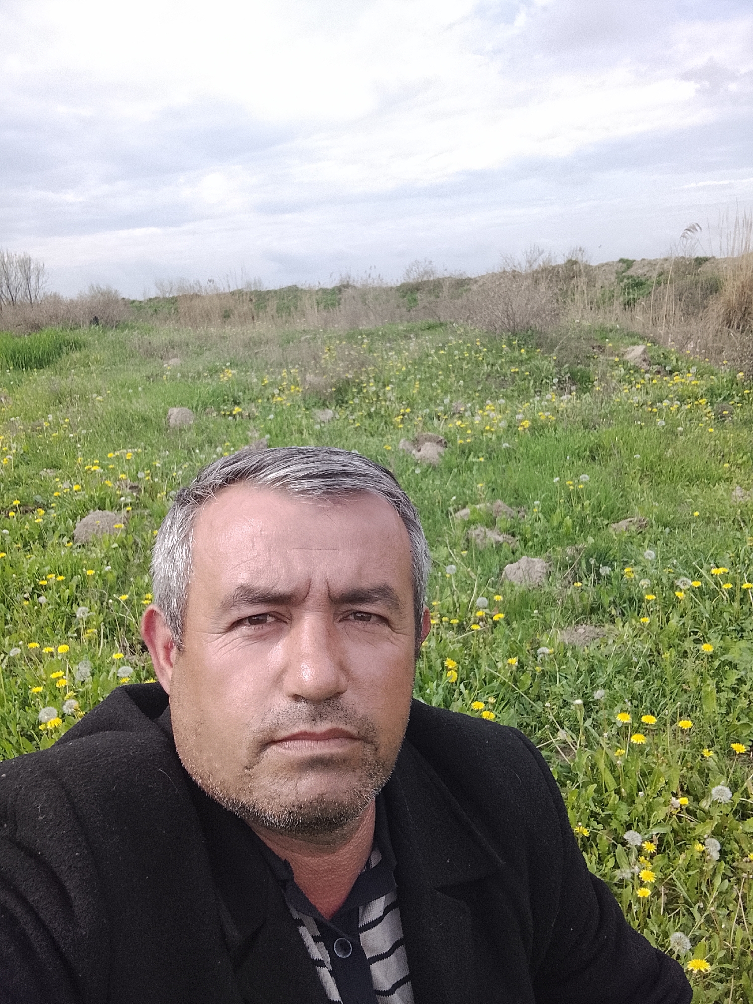 A man is in a field with green grass and yellow flowers under a cloudy sky. The man has gray hair and stubble on his face. He is wearing a black coat over a striped shirt. The background includes vegetation and possibly a small hill or mound. The location may be Sabir Rakhimov, Uzbekistan.