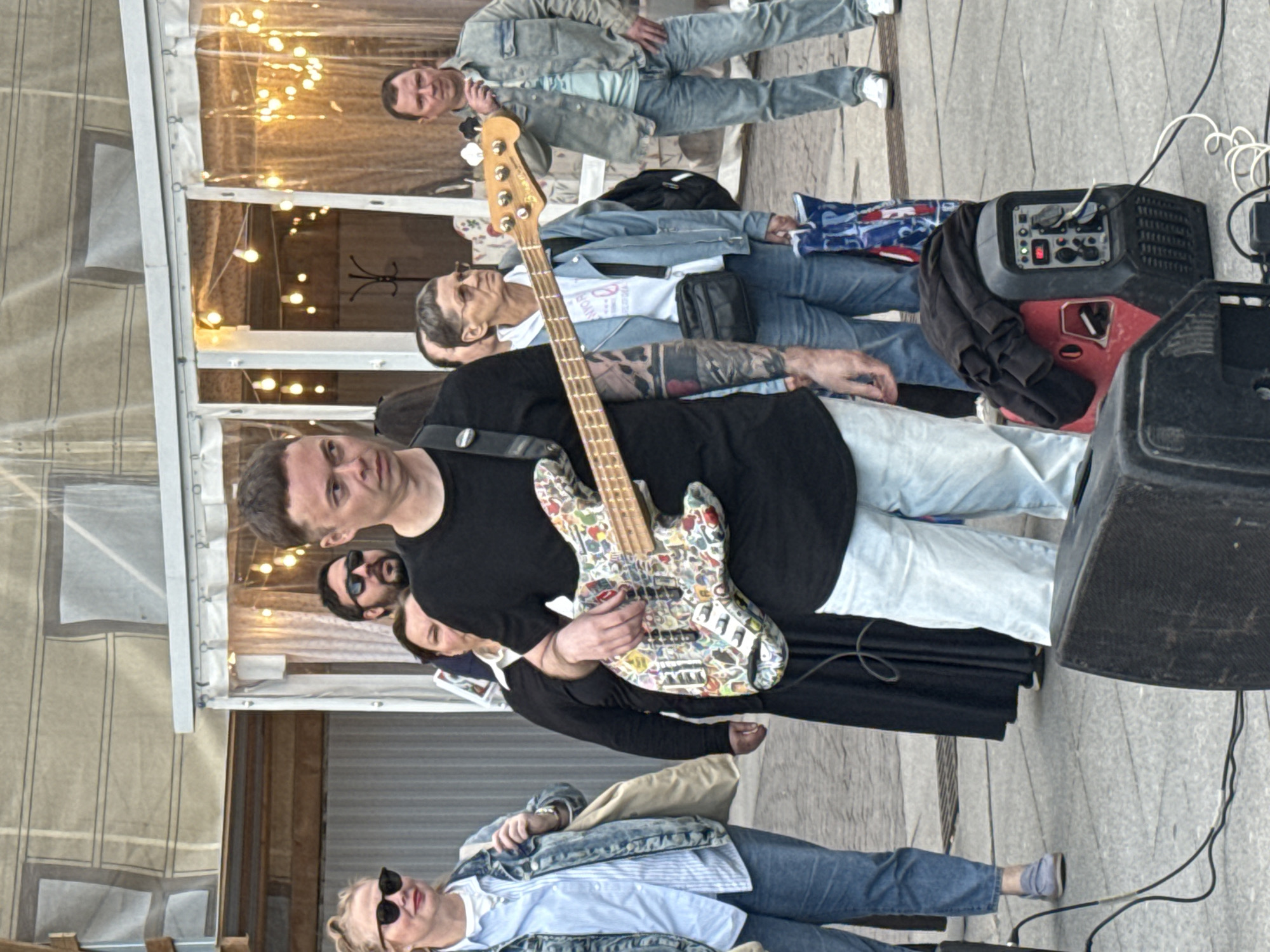 The image depicts an outdoor scene, likely a performance or public gathering in Saint Petersburg, Russia. In the foreground, a male musician is playing a bass guitar adorned with numerous colorful stickers. He is wearing a black t-shirt and light-colored pants. To his left, a woman in a denim jacket and blue jeans stands looking towards the viewer. Behind the musician, several other individuals are visible, including a man with sunglasses and another woman. In the background, a transparent tent structure with string lights is present. The ground is paved with grey square tiles. A red and black generator and a speaker are positioned in the lower right corner of the frame, connected by cables. The overall atmosphere suggests a casual, outdoor event.