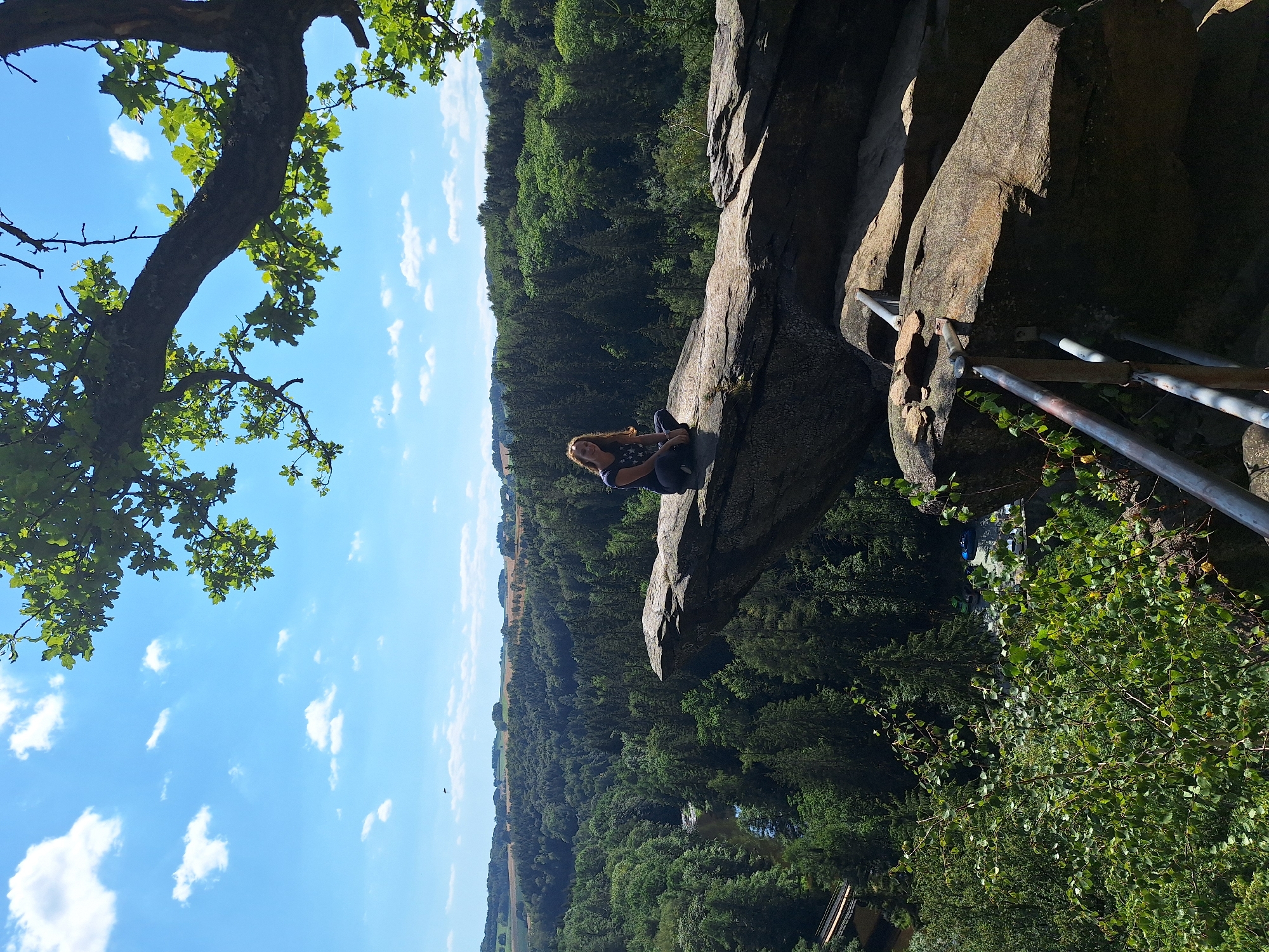 A woman is pictured sitting cross-legged on a prominent rock formation overlooking a lush, forested valley. The rock she sits on juts out dramatically, offering a precarious vantage point. The valley below is densely populated with trees, with a river winding through it. In the distance, rolling hills and fields suggest a rural landscape. The sky is blue with scattered white clouds. A large tree branch with green leaves frames the top right of the image. The scene appears to be in the vicinity of Wolkenstein, Germany, known for its scenic rock formations and forests, likely a hiking or natural attraction area.