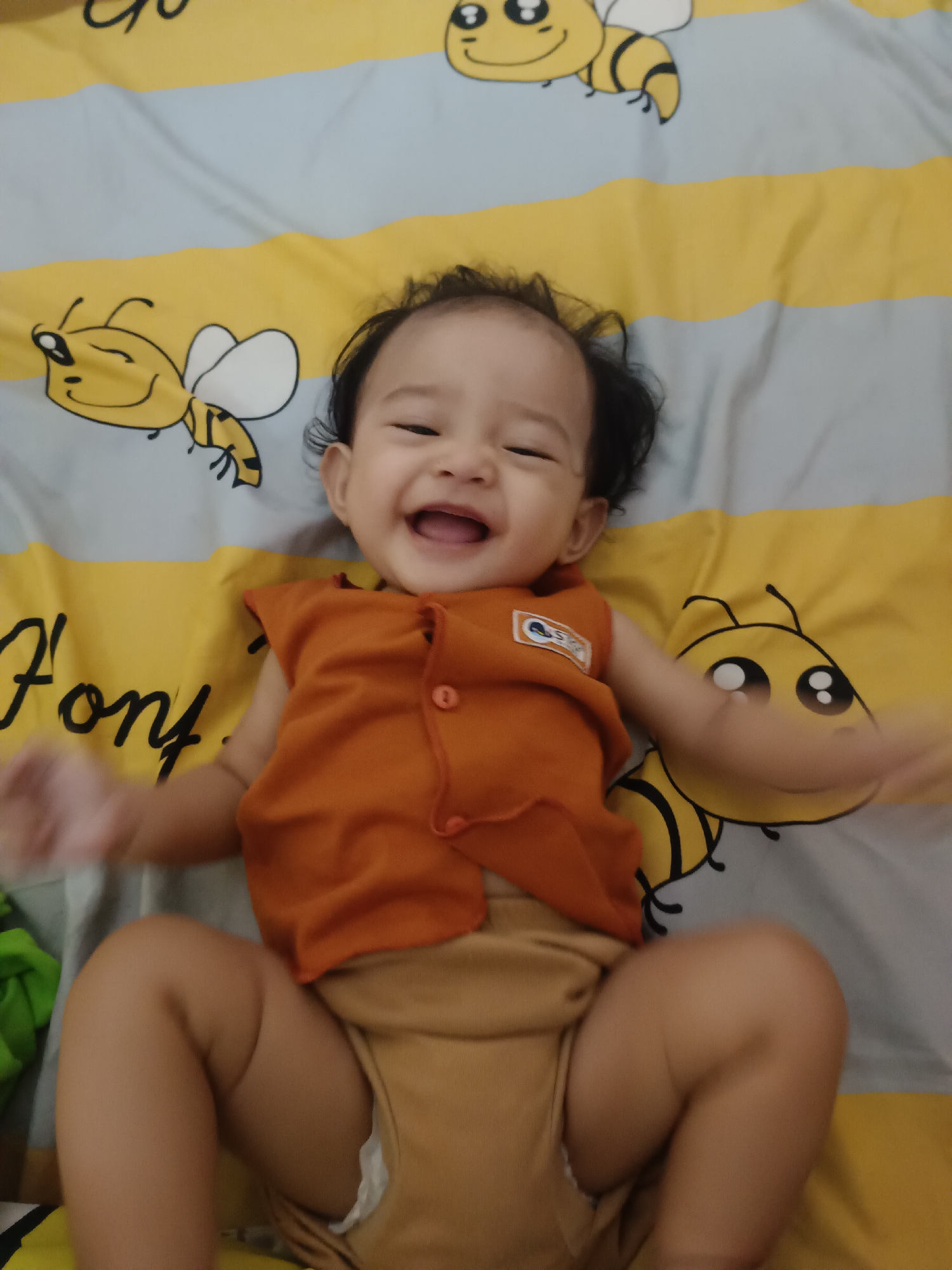 A baby is lying on its back on a yellow and grey striped sheet with bee illustrations. The baby is wearing an orange sleeveless shirt and a tan diaper. The baby is smiling and appears to be laughing. The scene is likely indoors, possibly in a bedroom or nursery. There is no indication of the location being in the city of Kawua, Indonesia.