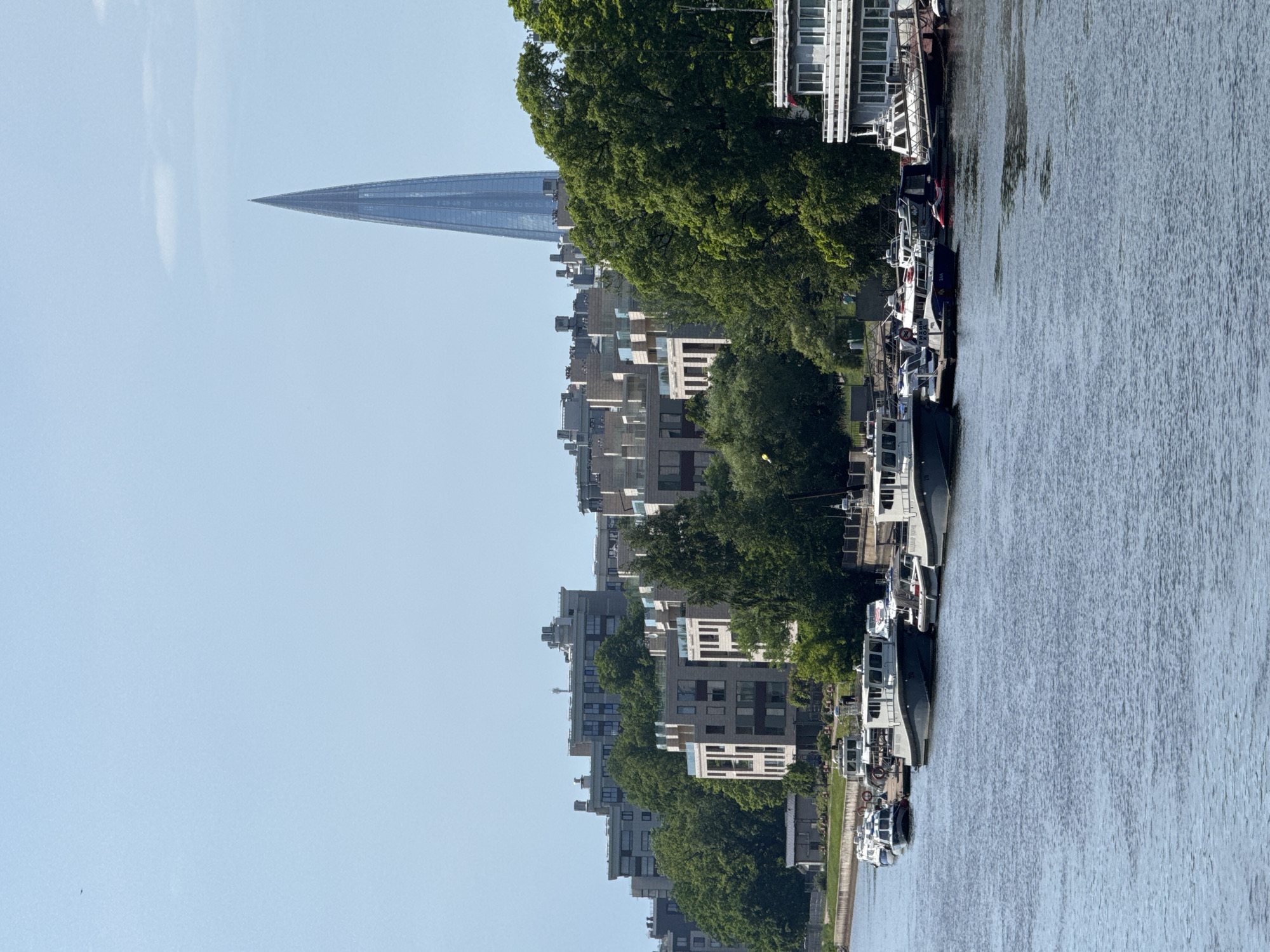 This image shows a waterfront scene in Saint Petersburg, Russia, likely along the Neva River or one of its tributaries. The foreground is dominated by the calm, blue water of the river. Moored along the left bank are several boats of varying sizes, including what appear to be passenger vessels and smaller motorboats. Some of these boats are docked, while others are gently bobbing on the water.
Beyond the boats, a treeline of lush green trees separates the waterfront from a complex of modern, multi-story residential buildings. These buildings feature a contemporary architectural style with clean lines and balconies. In the background, a prominent, needle-shaped skyscraper, likely the Lakhta Center, pierces the clear blue sky, its glass facade reflecting the daylight. The overall impression is one of a well-developed urban waterfront area with a blend of residential development and modern architecture. There are no visible people or specific actions taking place in the frame.