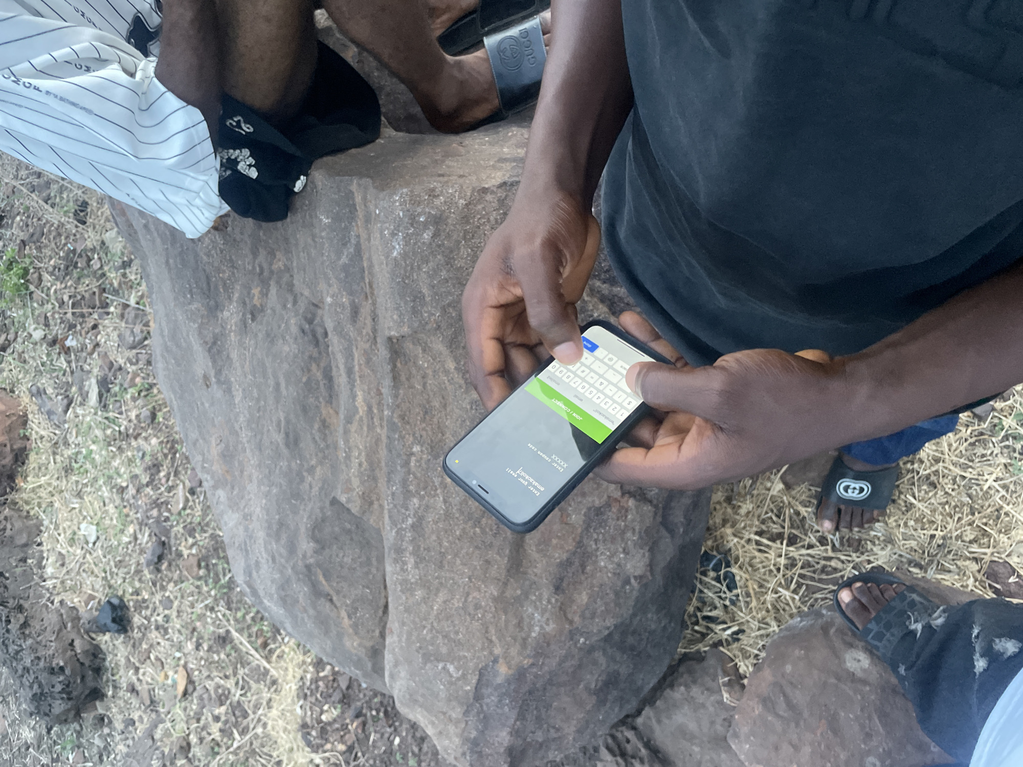 This overhead shot captures a casual outdoor scene set among large, grey-brown rock formations, likely in Lokoja, Nigeria, as per the provided context. The ground is sparsely covered with dry grass and loose dirt, characteristic of a natural or undeveloped area.
The primary subject is a person, whose dark-skinned hands are actively engaging with a black smartphone. The phone screen displays a digital form with text fields, including "Enter your email" (with "emakaokoli" visible, possibly a pre-filled or typed entry), "Enter coupon code" (showing "XXXXXX"), and a prominent green "JOIN / CONNECT" button. A standard numeric keyboard (1234567890) is visible at the bottom of the screen, along with the word "emailed" partially visible near the top right. This suggests the individual is completing an online registration or form.
Visible around the person holding the phone are parts of two other individuals. To the upper left, a person is seen wearing white striped shorts or pants, featuring partial text "CMGF A BATHING APE." Black socks worn by this individual display some shiny embellishments and the number "23." To the lower right, another person's feet are visible, sporting black slides, one of which is clearly branded with the "GUCCI" logo. The scene is brightly lit by natural daylight, indicating a time around midday or early afternoon. The overall ambiance appears relaxed and informal.