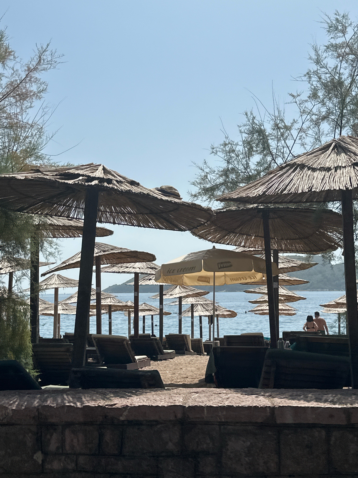 The image depicts a beach scene on a sunny day in Budva, Montenegro. In the foreground, a stone wall lines the bottom of the frame. Behind the wall, a sandy beach is visible with rows of empty lounge chairs and straw umbrellas. The umbrellas are predominantly made of natural materials, casting shadows on the sand. The sea is calm and blue, with a few distant islands or hills visible in the background. A couple, a man and a woman, are seen walking towards the water on the right side of the image. The trees with sparse foliage frame the top portion of the scene, suggesting a coastal environment. The overall atmosphere is one of a relaxed beach resort.