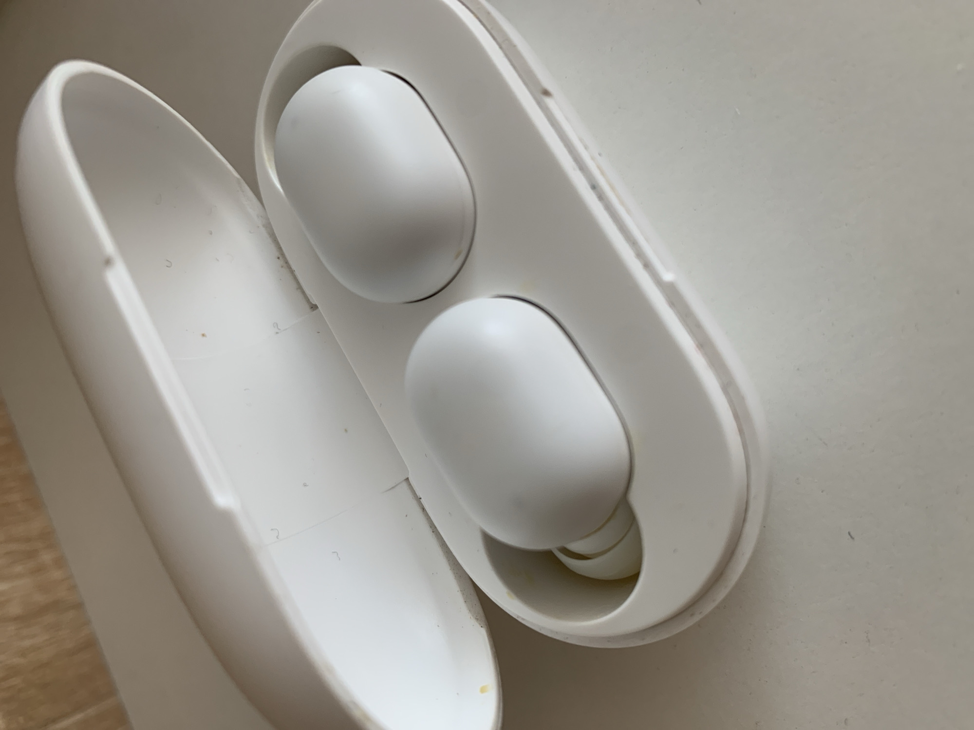 The image shows a white wireless earbud charging case with two white earbuds inside. The case is open, revealing the earbuds nestled in their charging slots. The earbuds are oval-shaped and appear to be made of plastic. The charging case also appears to be made of plastic and has a smooth, matte finish. The earbuds are positioned for charging, with the left earbud closer to the viewer and the right earbud slightly further away. The charging case lid is open and tilted back, resting against the surface it is placed on. The surface appears to be a light-colored, textured material, possibly a desk or table. There are a few small specks of dust or lint visible on the inside of the charging case lid and on the earbuds themselves. The lighting suggests an indoor environment, possibly with natural light from a window. There is no visible indication of the location being in the city of Ternopil, Ukraine, or any specific geographical details.