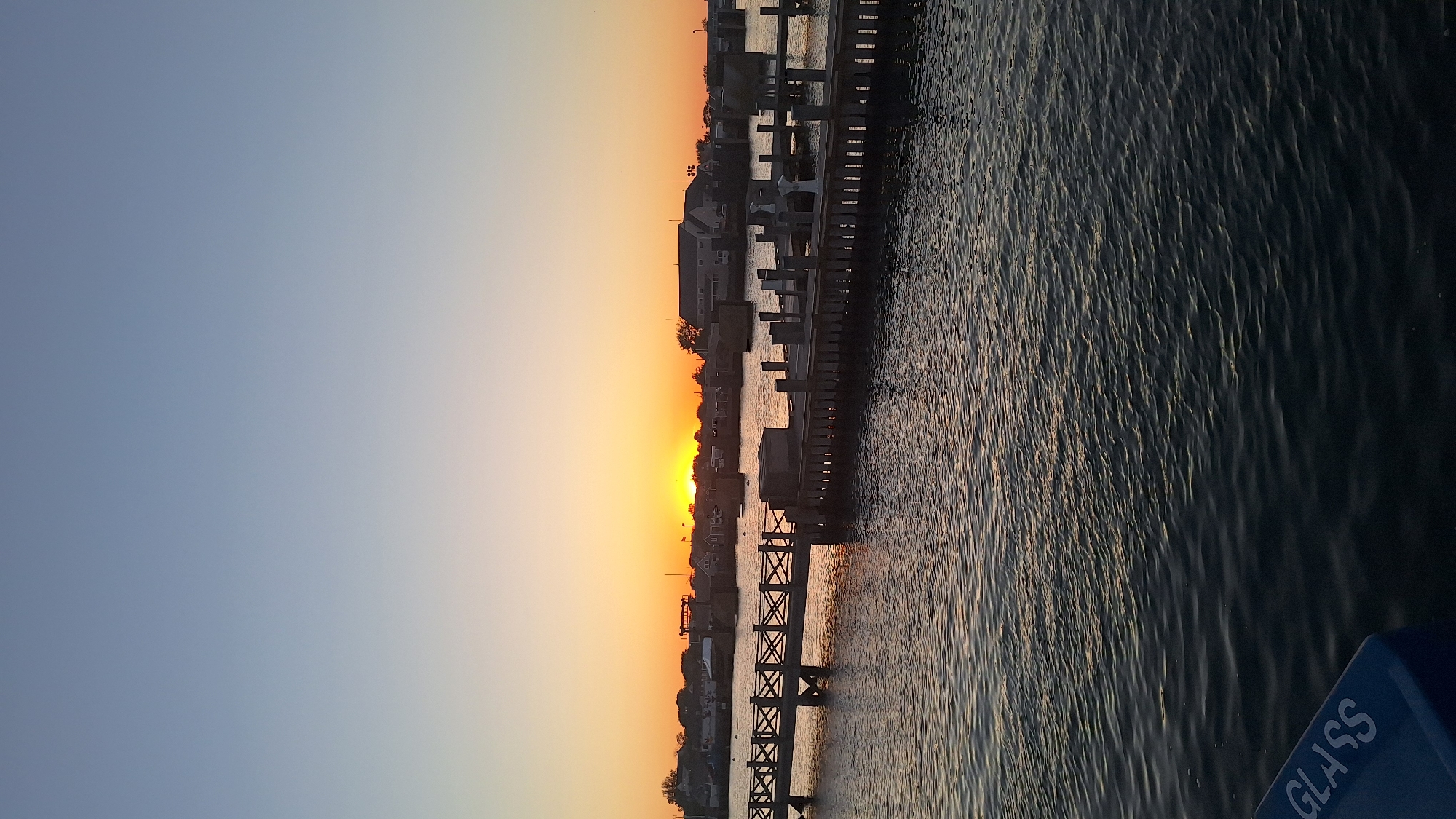 The image captures a serene sunset over a harbor, likely in Nantucket, United States of America. The sky transitions from a pale blue at the top to a vibrant orange glow near the horizon, where the sun is setting. A wooden boardwalk or pier stretches across the middle of the frame, with a series of pilings supporting it. Beyond the boardwalk, several buildings, possibly houses or marine businesses, are visible along the coastline, silhouetted against the colorful sky. The water in the foreground is dark and reflects the colors of the sunset, with gentle ripples distorting the reflections. In the lower left corner of the image, a blue recycling bin with the word "GLASS" printed on it is partially visible. No people are present in the scene.