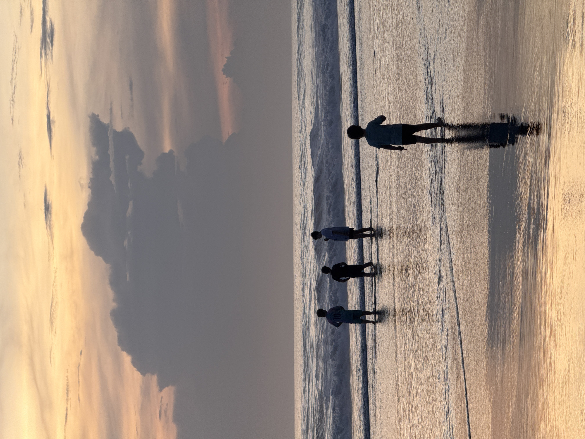 The image shows a beach scene at sunset in North Kuta, Indonesia. The sky is filled with clouds, ranging from light orange and yellow hues near the horizon to darker gray near the top of the image. In the foreground, there are four people walking along the wet sand near the water's edge. The people are silhouetted against the reflective surface of the beach, with the light source behind them. The receding waves are creating patterns on the sand. The people appear to be spaced out, with one further ahead of the other three.