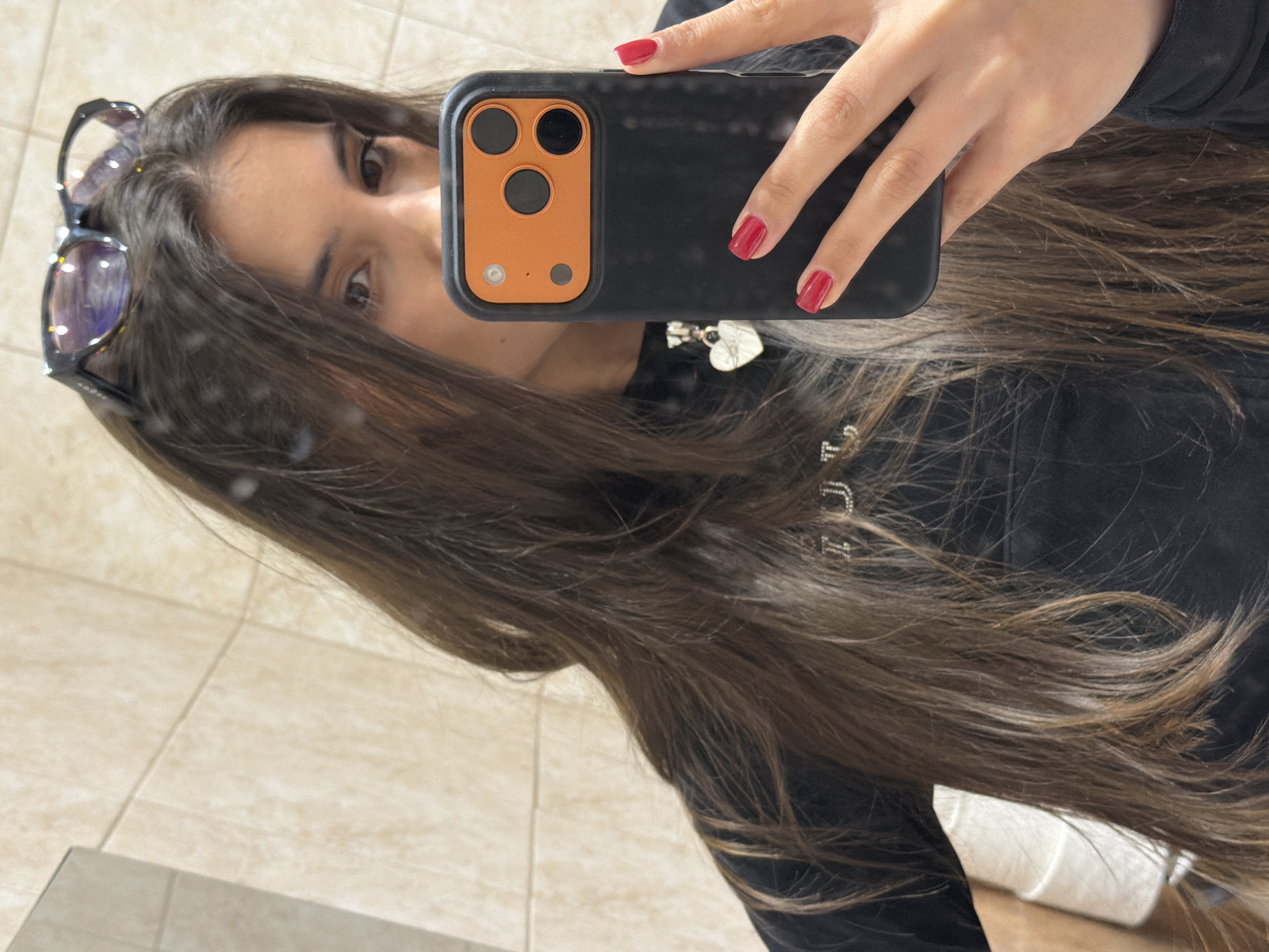 A young woman is taking a selfie in a bathroom. She is holding a black phone with an orange camera surround, featuring three lenses and a flash. The phone obscures part of her face, but her eyes, part of her nose, and mouth are visible. Her fingernails are painted red. She has long, brown hair. Sunglasses are perched on her head. She is wearing a black hooded sweatshirt, with a heart-shaped pendant visible around her neck. The background shows light beige tile and a glimpse of a toilet. The specific location in Sofia, Bulgaria cannot be determined from the image.