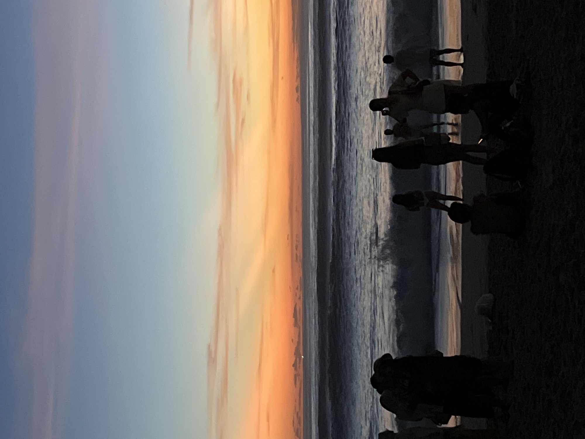 Here is a forensic description of the image:
The image depicts a beach scene at sunset, oriented vertically. In the foreground, on the right side, are silhouetted figures of people standing on the beach. Their forms are dark and not clearly defined. A large wave is crashing in the center of the image, visible between the sky and beach. The sky is a blend of blue and orange hues, typical of a sunset. There are indistinct cloud formations. The location is possibly in Kuta Selatan, Indonesia.