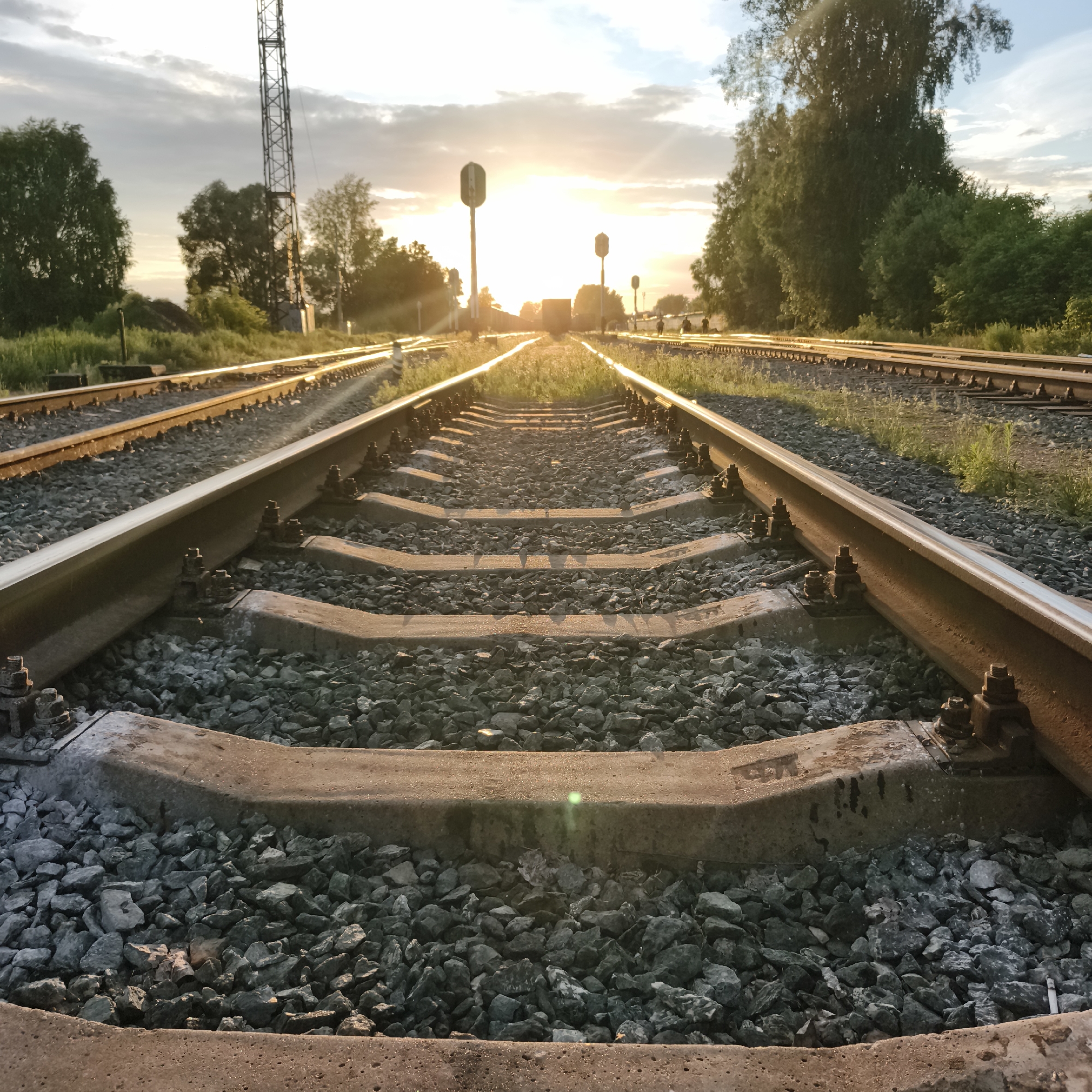 This media file depicts a railway track at sunset. The sun is low in the sky, casting a warm, golden light across the scene and creating lens flare effects. The tracks, made of steel rails and concrete sleepers, are surrounded by gravel ballast and green vegetation. Several railway signals are visible in the distance, with one illuminated. Trees line the sides of the track, some appearing to be birch trees, common in Russia. The sky is a mix of blue and white clouds, indicative of a partly cloudy day. The overall atmosphere is serene and tranquil, capturing the quiet beauty of the end of the day at a railway junction. There are no people visible in the frame. The location context is Chaikovsky, Russia, a city known for its industrial and railway infrastructure. The time of day is clearly sunset, and the weather appears calm. There is no visible text in the image.