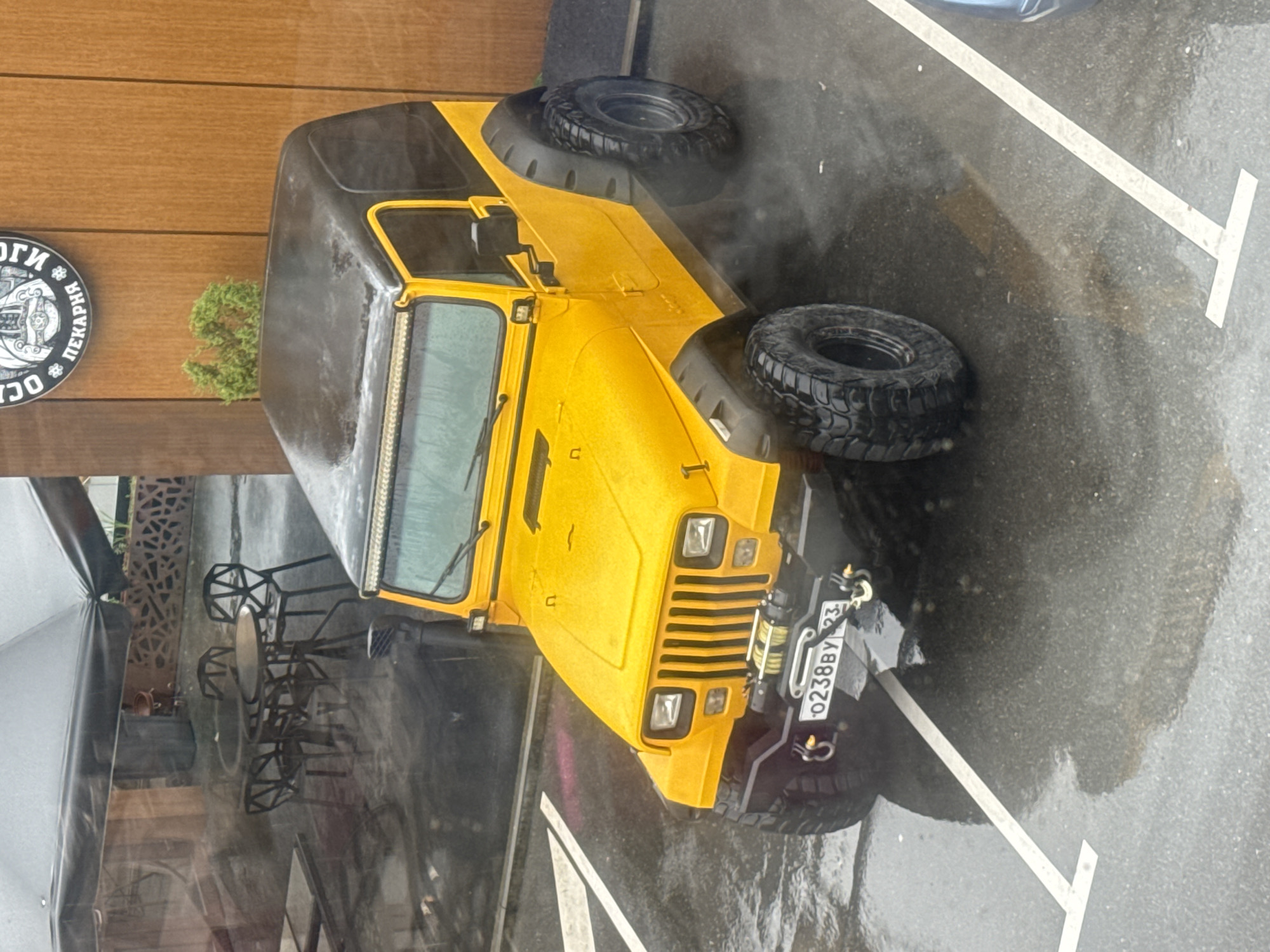 The image depicts a yellow and black Jeep parked in a designated parking space adjacent to a building. The parking space is marked with white lines on wet asphalt. The Jeep has the license plate number "O238BY 23". Seating and tables are visible beside the building. A sign on the building indicates the presence of a bakery, written in Cyrillic. Based on the Cyrillic text, the location is assumed to be Sochi, Russia. There is no evidence of people.