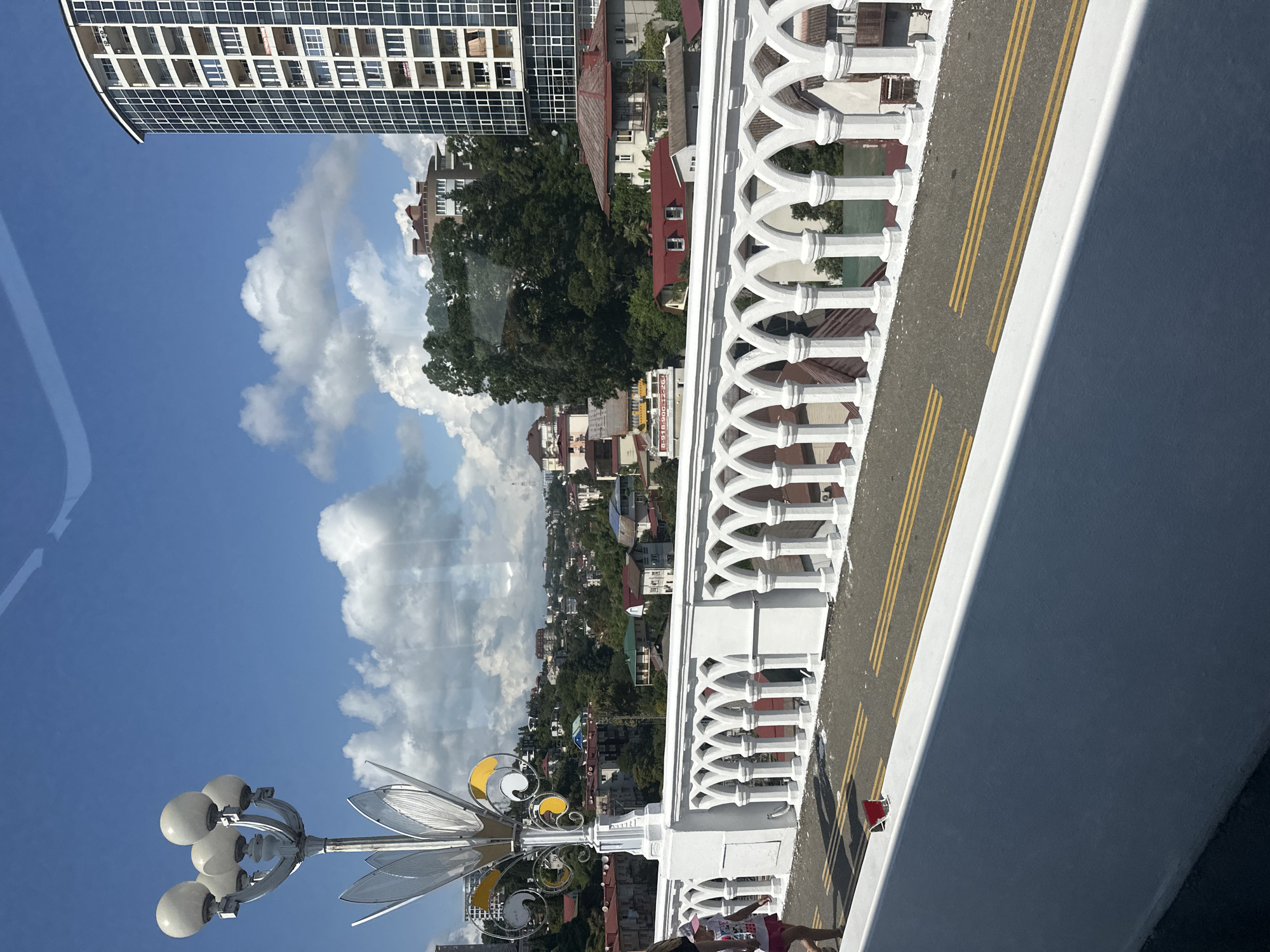 A wide shot shows a white ornate bridge with arched railings. Yellow double lines mark the road on the bridge. A child in a pink hat and shorts walks on the left side of the bridge. In the background, a cityscape of houses and buildings is visible on a hill, with a tall, modern, glass-enclosed skyscraper to the right. A street lamp with multiple bulbs is on the left side of the bridge. The sky is blue with white clouds. The scene appears to be in Sochi, Russia, based on the architecture and the distinctive curved skyscraper, which resembles buildings found in the city's coastal areas.