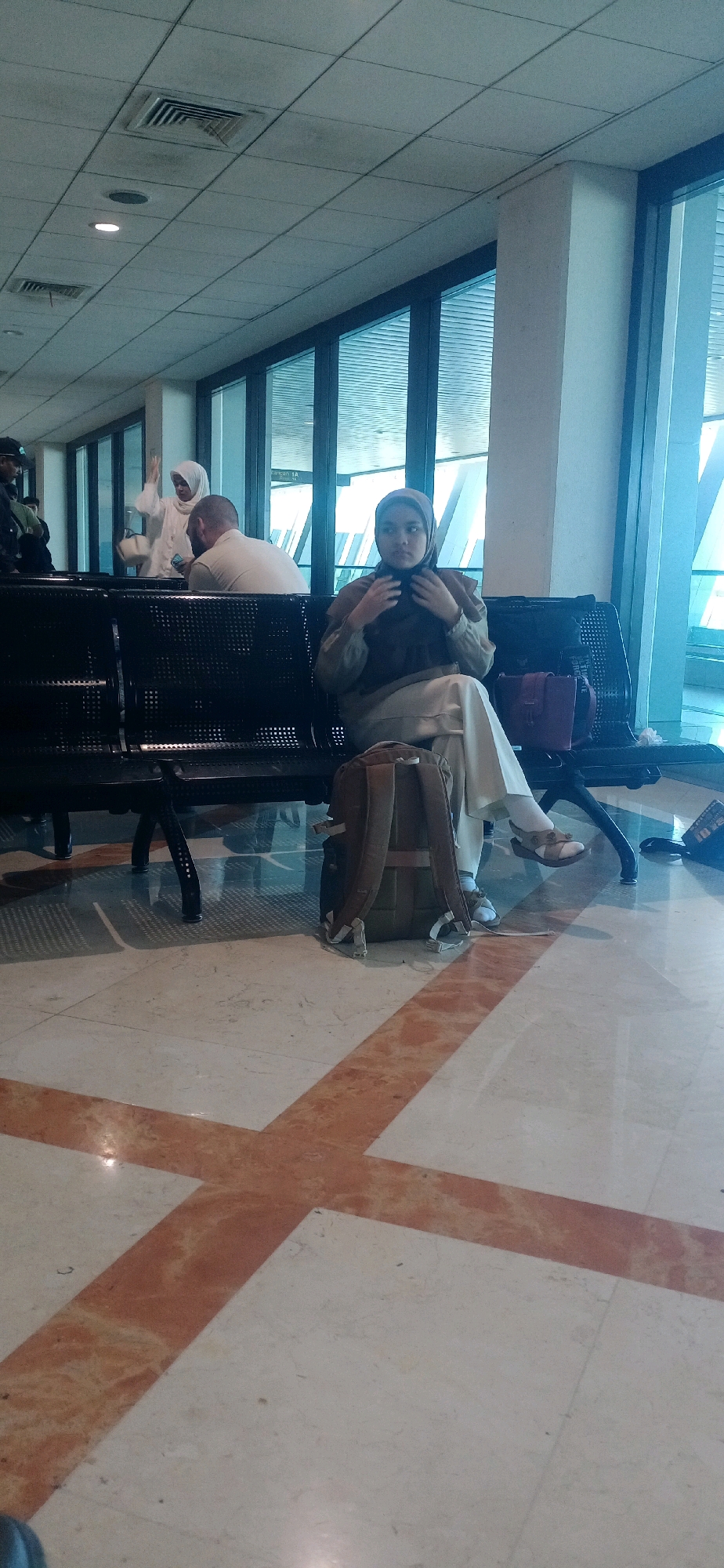 The image depicts an interior scene, likely an airport waiting area, with a young woman wearing a hijab and loose-fitting clothing sitting on a dark bench. She is holding her hands to her neck. A backpack is on the floor next to her, and another bag is on the bench beside her. In the background, other individuals are visible, including a woman in a white hijab and a bald man. The environment features large windows with a blue tint, a tiled floor with marble and red accent strips, and a suspended ceiling with fluorescent lighting and ventilation grilles. The scene is indoors and appears to be during the day, with natural light coming through the windows. The location is specified as Segoro Tambak, Indonesia.