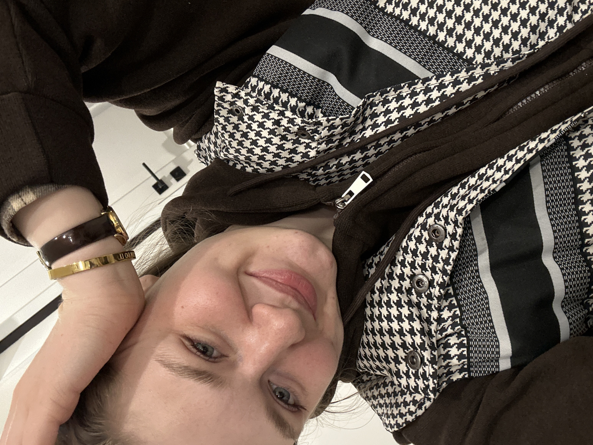 A single female individual is depicted in a close-up, upward-angled indoor shot. She has a light complexion, light-colored eyes, and light brown hair partially visible. Her facial expression is a closed-mouth smile, with noticeable rosy cheeks and light pink lipstick. She is wearing a dark brown hooded jacket, unzipped, over which a black and white houndstooth patterned garment is visible. This garment also features horizontal black and grey stripes with fine dot patterns. On her right wrist, she wears a gold-colored bangle bracelet inscribed with "JUST DO IT" and a dark, wide-strapped watch with a gold-colored buckle. Her right hand is raised, with the wrist resting against the top of her head/forehead. The background consists of a white wall, displaying two black, minimalist door handles or similar fixtures on a white surface in the upper right quadrant. The location is an indoor setting, but no specific visual cues confirm a precise location within the city of Kazan, Russia.
