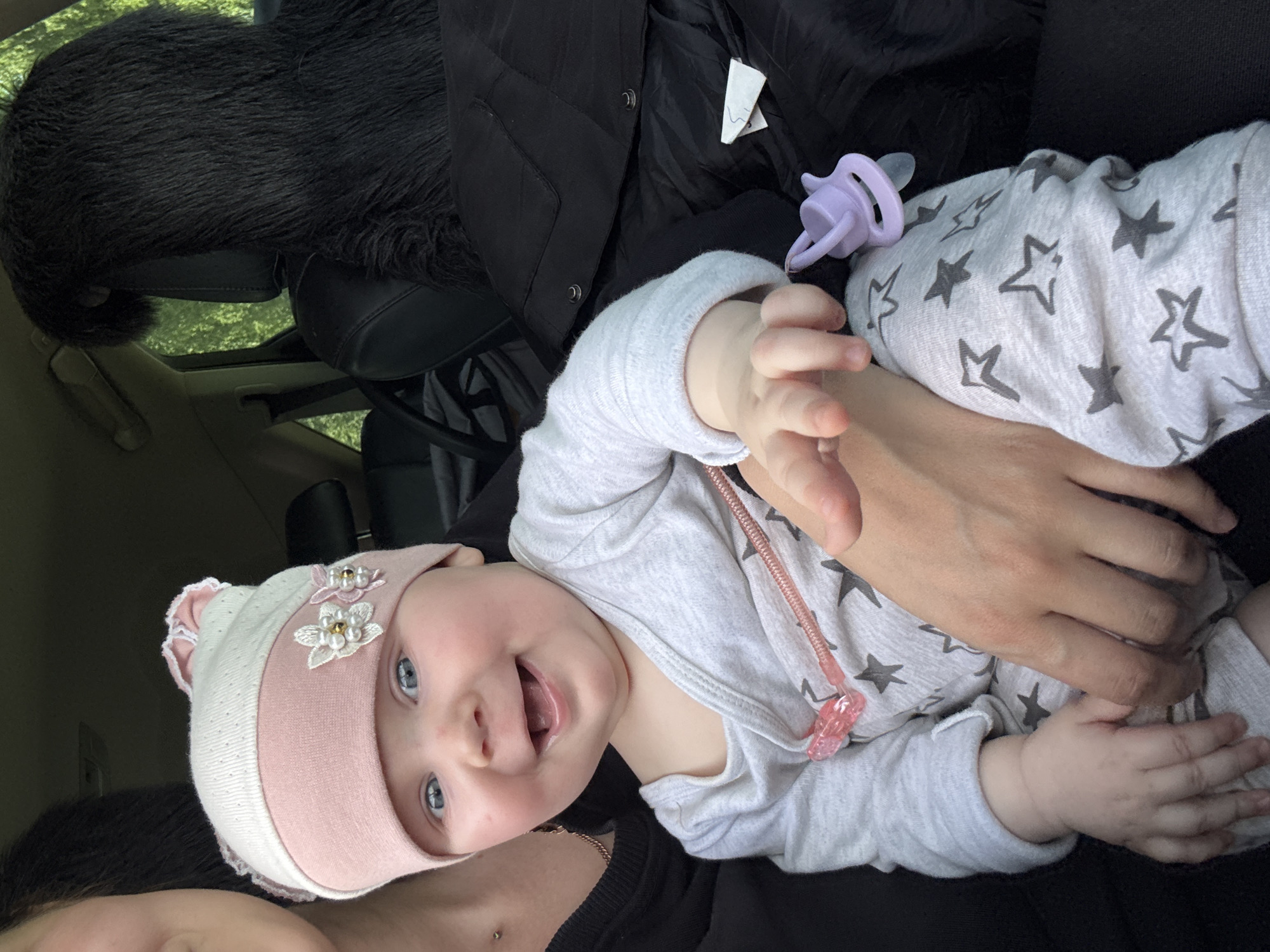 A baby, lying on their back and wearing a light gray onesie with dark gray stars, is being held. The baby is smiling and has a light pink and white hat with decorative flowers on. A lavender pacifier is near the baby. The environment appears to be inside a car, with a black fluffy headrest visible in the background. The location cannot be identified in the city of Votkinsk, Russia.