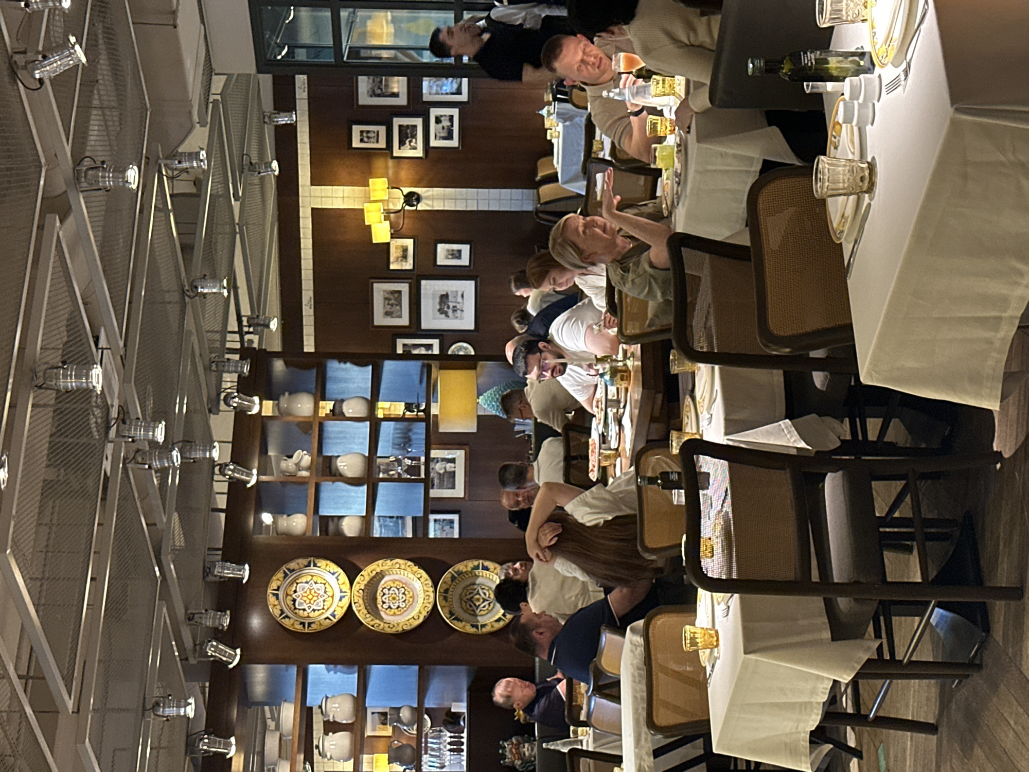 The image depicts the interior of a restaurant, likely in Moscow, Russia, with several tables set for dining and occupied by patrons. The decor features a rustic and warm ambiance, with wooden accents, framed pictures on the walls, and decorative plates displayed on shelves. Spotlights hang from a meshed ceiling, illuminating the dining area.
In the foreground, a table is set with a white tablecloth, plates, cutlery, glasses, and a bottle of olive oil. Two chairs with woven backs are positioned at the table. Further back, other tables are visible, some with diners engaged in conversation or eating. People of various ages are seated at the tables, dressed in casual attire.
The restaurant appears to be busy, with staff members visible in the background, including one man in a black shirt who seems to be attending to guests. The overall atmosphere suggests a lively and social dining experience. The presence of Italian-style decorative plates on the shelves might hint at the restaurant's cuisine or theme.