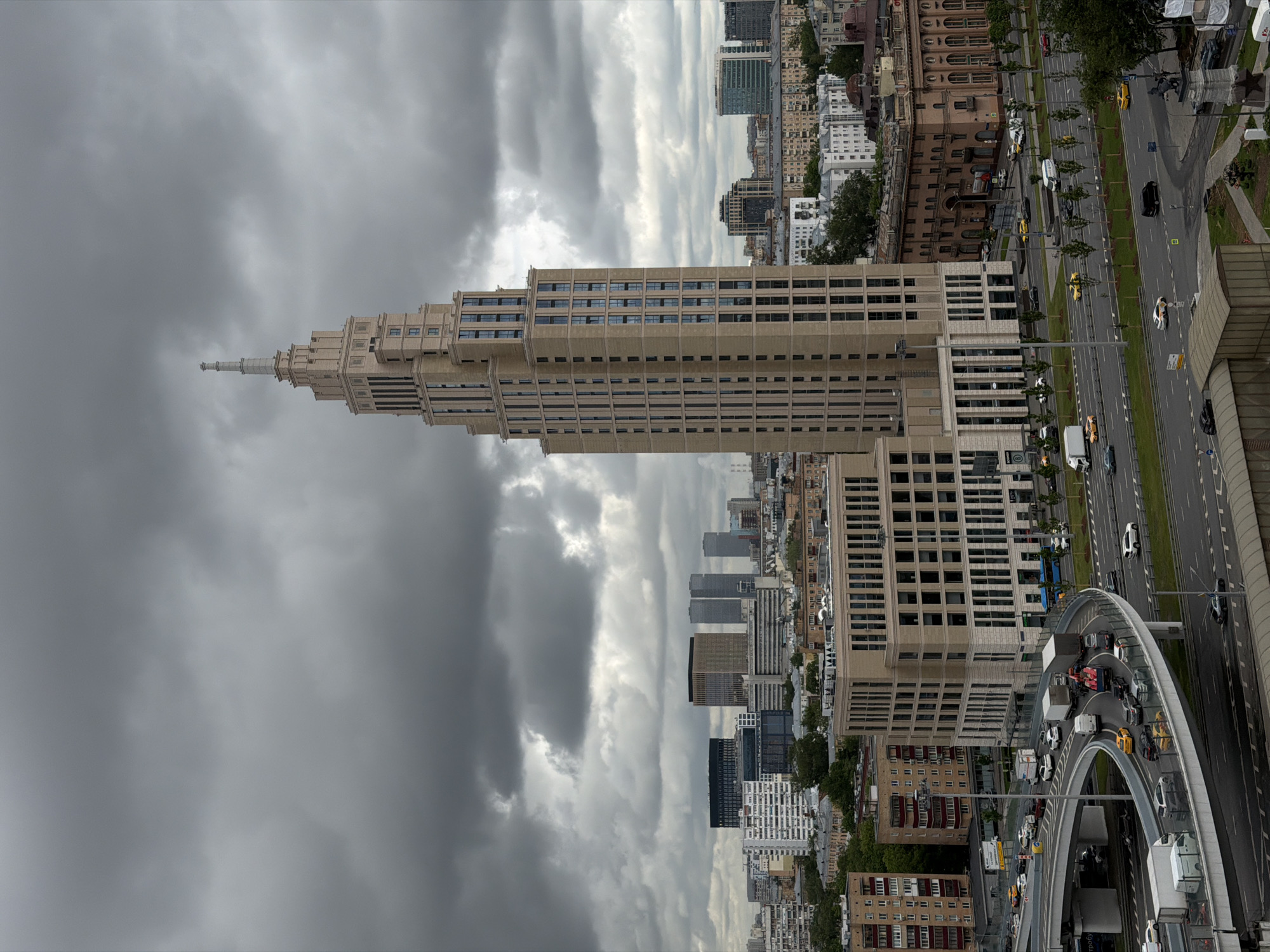 The image shows a view of Moscow, Russia, featuring the Triumph Palace skyscraper prominently. The building is tan with a stepped, pyramidal top and a slender spire. Dark storm clouds fill the sky behind and to the left of the building.
To the right of the skyscraper, a busy roadway stretches into the distance, lined with trees and a mix of low-rise and taller buildings. Traffic is visible on the road, including cars, vans, and taxis. A curved overpass intersects the roadway, also carrying traffic.
The scene conveys a contrast between the modern architecture of the skyscraper and the older buildings in the city. The overcast sky contributes to a somewhat somber atmosphere.