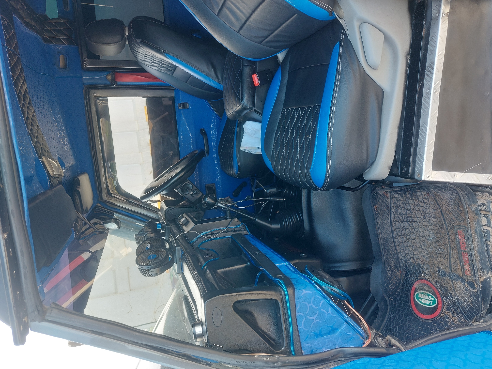 The image shows the interior of a vehicle, specifically the driver's side. The vehicle has black and blue seats, a steering wheel, a dashboard, and a floor mat with a Land Rover logo. The interior is primarily blue. The location is not discernible from the image alone, thus I cannot confirm it is Dar es-Salaam, Tanzania.