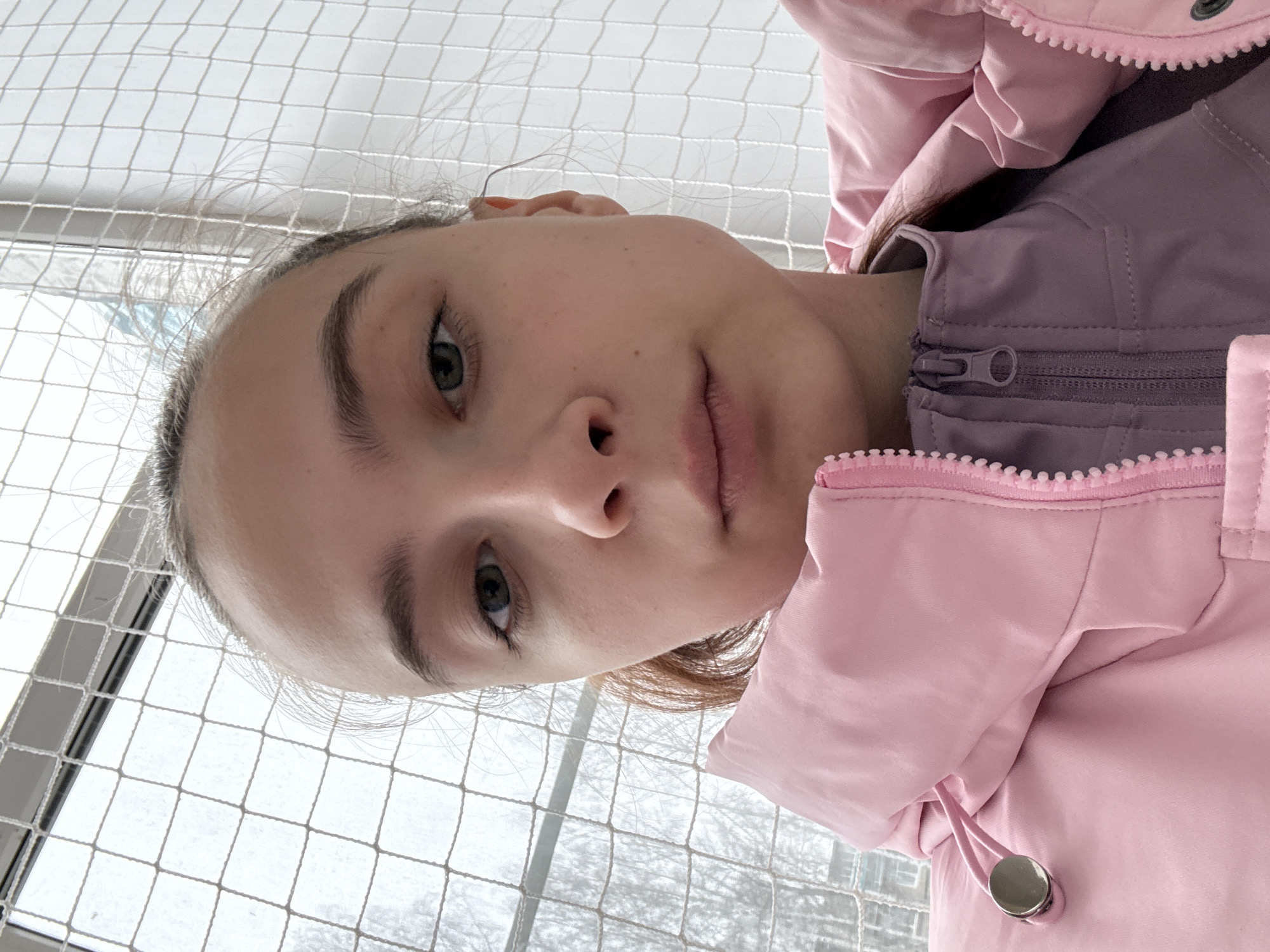 A young woman with fair skin and gray-green eyes is facing the camera from a low angle. She has light brown hair that is pulled back, and her facial expression is neutral. She is wearing a pink puffy jacket over a lavender zippered garment. Behind her is a white net-like structure with windows and trees visible in the background. The location is Chaikovsky, Russia.