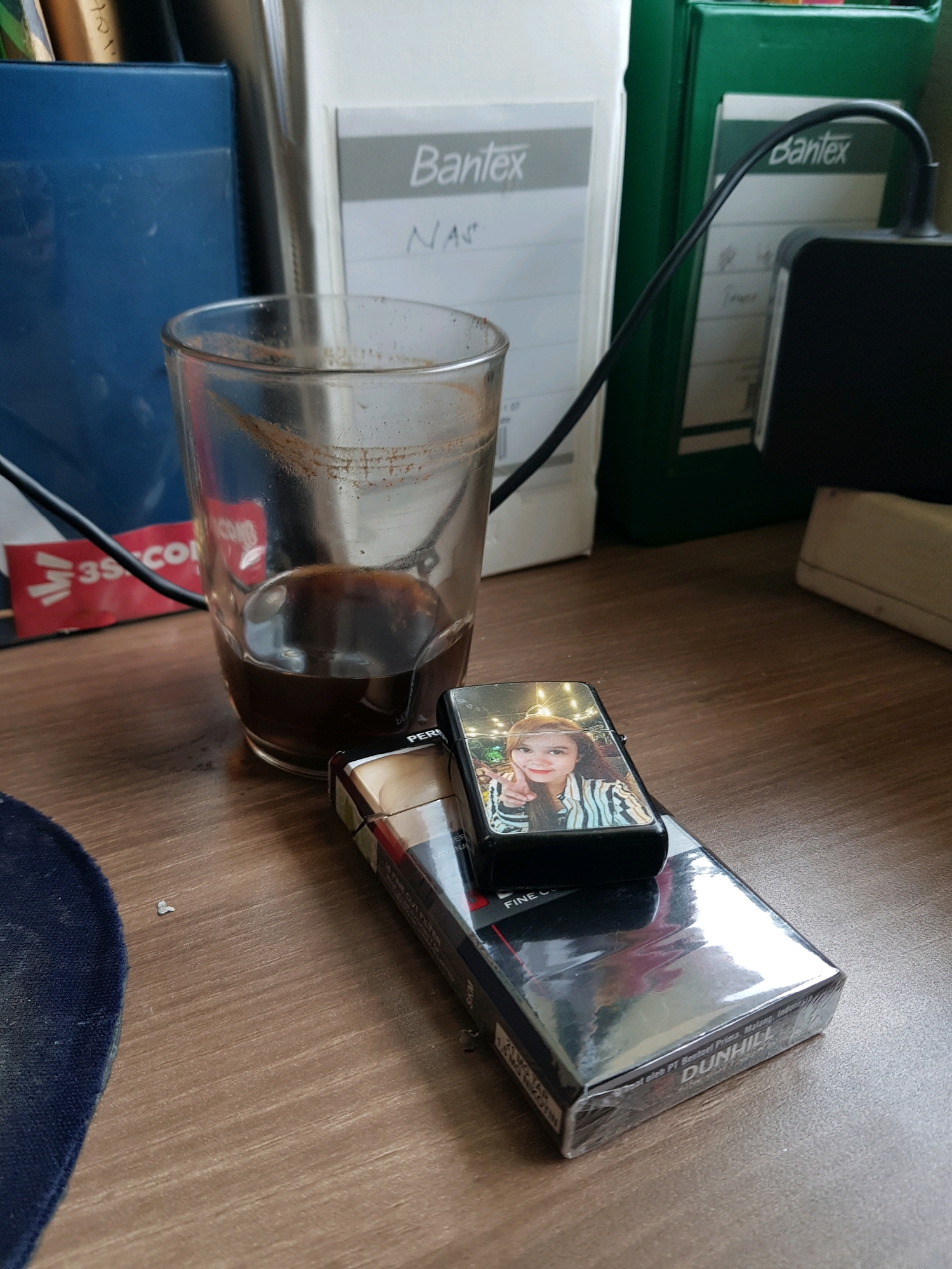The image shows a close-up of a wooden desk. In the foreground, a pack of Dunhill cigarettes is visible, partially wrapped in plastic. On top of the cigarette pack rests a black lighter with a picture of a young woman on it. To the left of the cigarettes is a glass containing a dark liquid, likely coffee, with some residue on the inside of the glass. In the background, various office supplies are arranged, including a blue container, a white binder labeled "Bantex," and a green binder. A black cable is also visible, connecting to an electronic device. The lighting suggests an indoor environment, possibly an office or home setting. The image does not provide any specific details about the location within Central Jakarta, Indonesia, or any actions or interactions occurring.