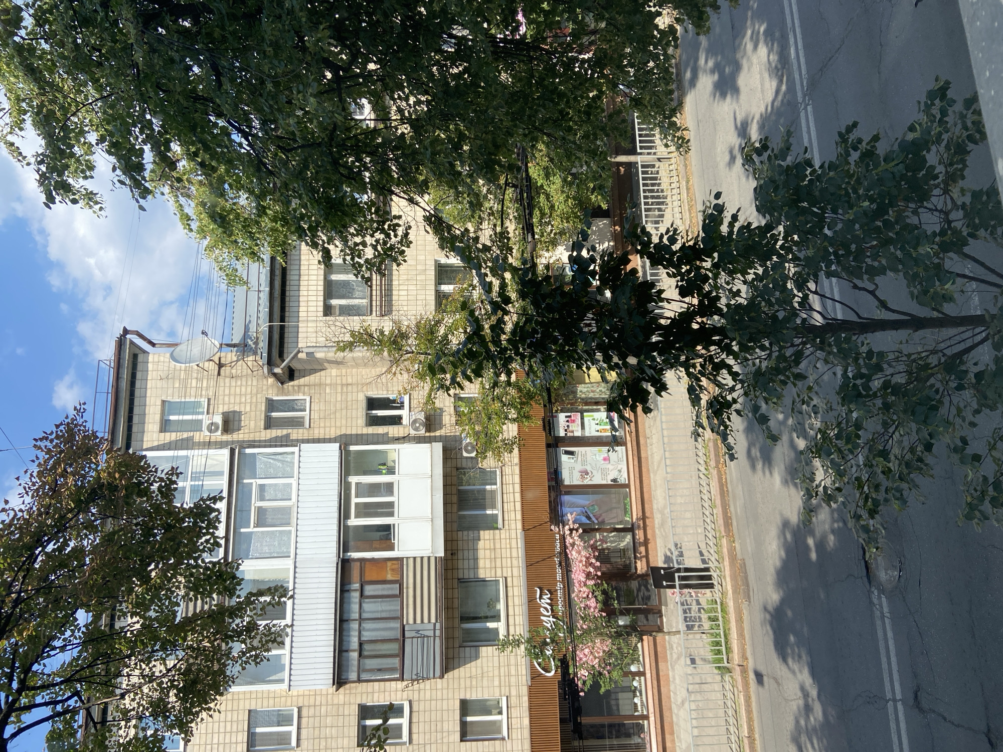The image depicts a street scene in Poltava, Ukraine, likely taken during daylight hours. In the foreground, a portion of an asphalt road with visible cracks and white lane markings is visible, with shadows cast by trees on its surface. A metal fence separates the road from a sidewalk, behind which a building with a storefront is situated. The building appears to be a multi-story structure with a beige facade. The storefront has large windows displaying various products, suggesting a retail establishment, possibly a beauty salon or a shop selling cosmetics, as indicated by text and product arrangements within the window. Above the storefront, the building has multiple windows and balconies on the upper floors. Several trees with green foliage are present in the frame, partially obscuring the view of the building and the sky, which is blue with some clouds. The overall impression is a typical urban environment in Poltava.