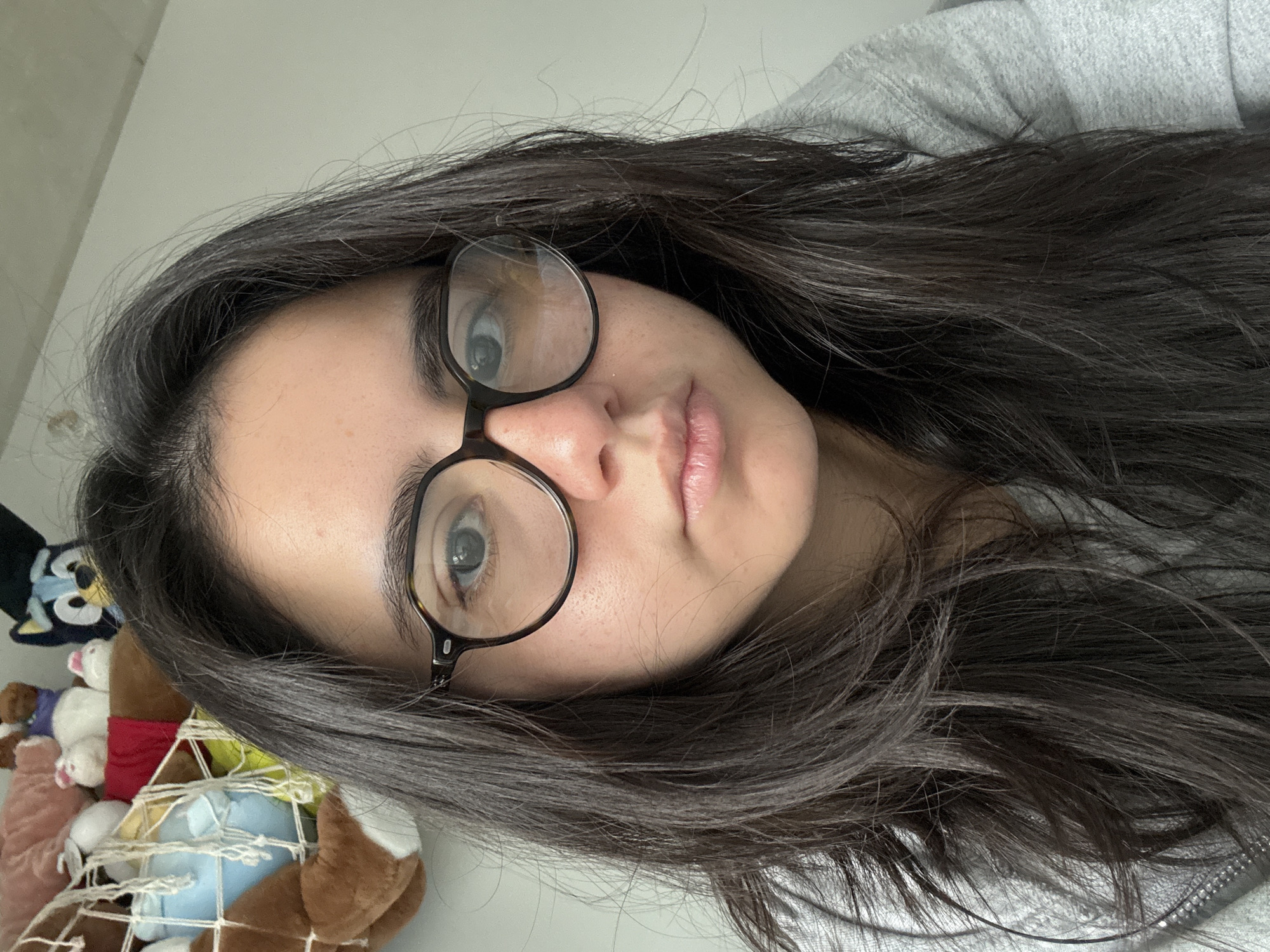 A young woman with long, dark hair and round glasses is positioned in the center of the frame, close to the camera. Her face is angled slightly to the right. The woman is wearing a gray top. To the left of the woman's face, there is a collection of stuffed animals held in a net. The background is a plain wall, partially visible. There is no information to identify the location as Austin, United States of America.