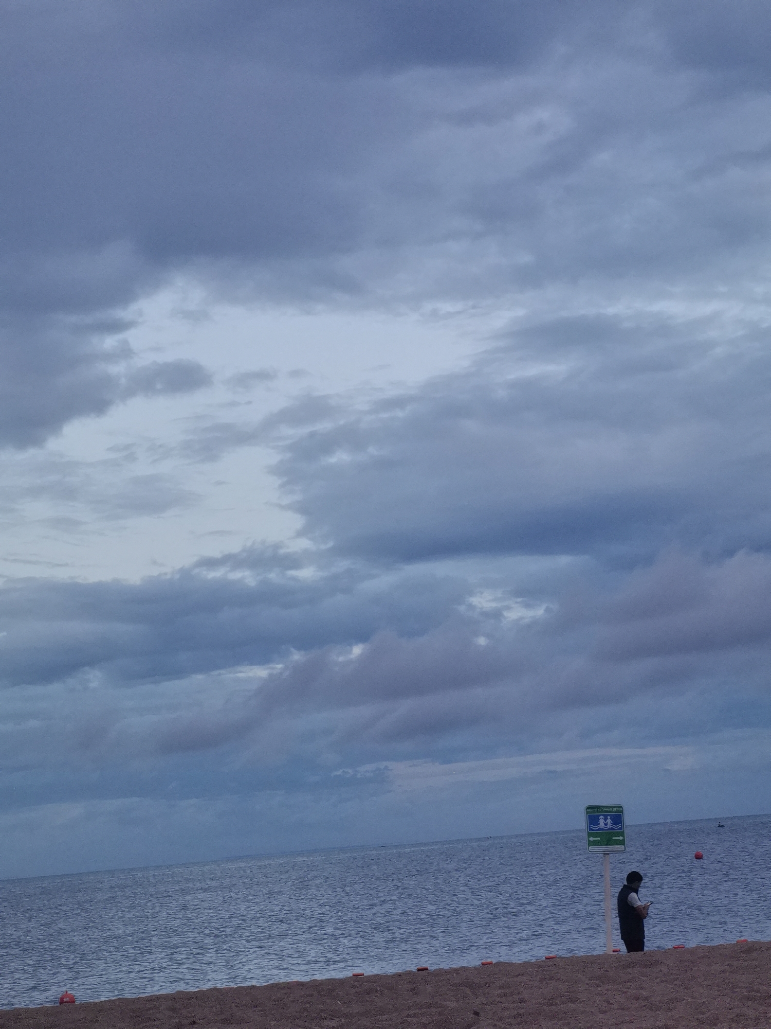 The image depicts a beach scene on a cloudy day. In the foreground, there is a sandy beach with two orange buoys visible. The middle ground features a calm body of water, likely the Gulf of Finland, given the location of Zelenogorsk, Russia. A person is standing on the beach near a green signpost. The sign has two blue figures swimming and two green arrows pointing in opposite directions, indicating swimming areas. The person, dressed in a dark vest over a light shirt and dark pants, is holding a mobile device and appears to be looking at it. Further out in the water, another orange buoy is visible. The background is dominated by a vast expanse of cloudy sky. The overall atmosphere is subdued and tranquil.