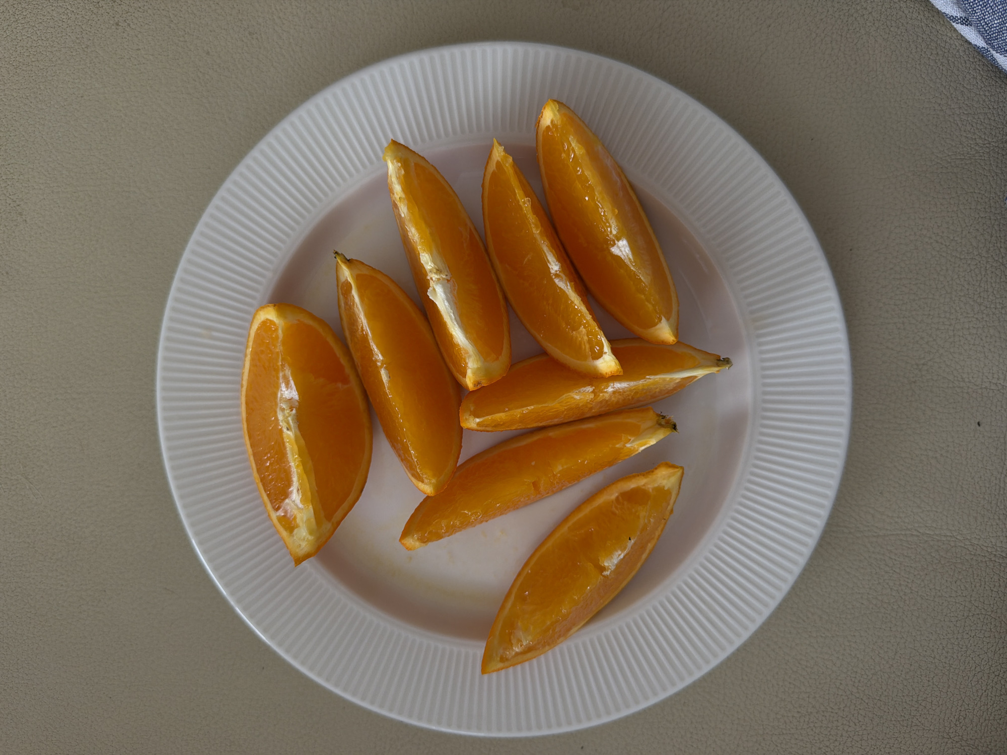 An overhead, close-up shot captures a white, circular plate filled with orange segments. Approximately ten to twelve freshly cut orange wedges are scattered on the plate, their vibrant orange flesh appearing juicy and slightly glistening. Some segments still show a thin line of white pith and traces of the outer orange peel.
The plate features a distinct design with radial ridges running along its wide rim. It rests on a textured, light beige or tan surface, which could be leather or a similar material, covering the majority of the background. In the top right corner of the frame, a small section of a blue and white striped fabric, likely a napkin or dishcloth, is partially visible.
The scene depicts a simple, prepared snack or part of a meal. No individuals are present, nor are there any discernible activities taking place beyond the presentation of the fruit. The image offers no visual cues regarding the time of day, weather conditions, or specific location within Budva, Montenegro, other than the provided context. There is no visible text.