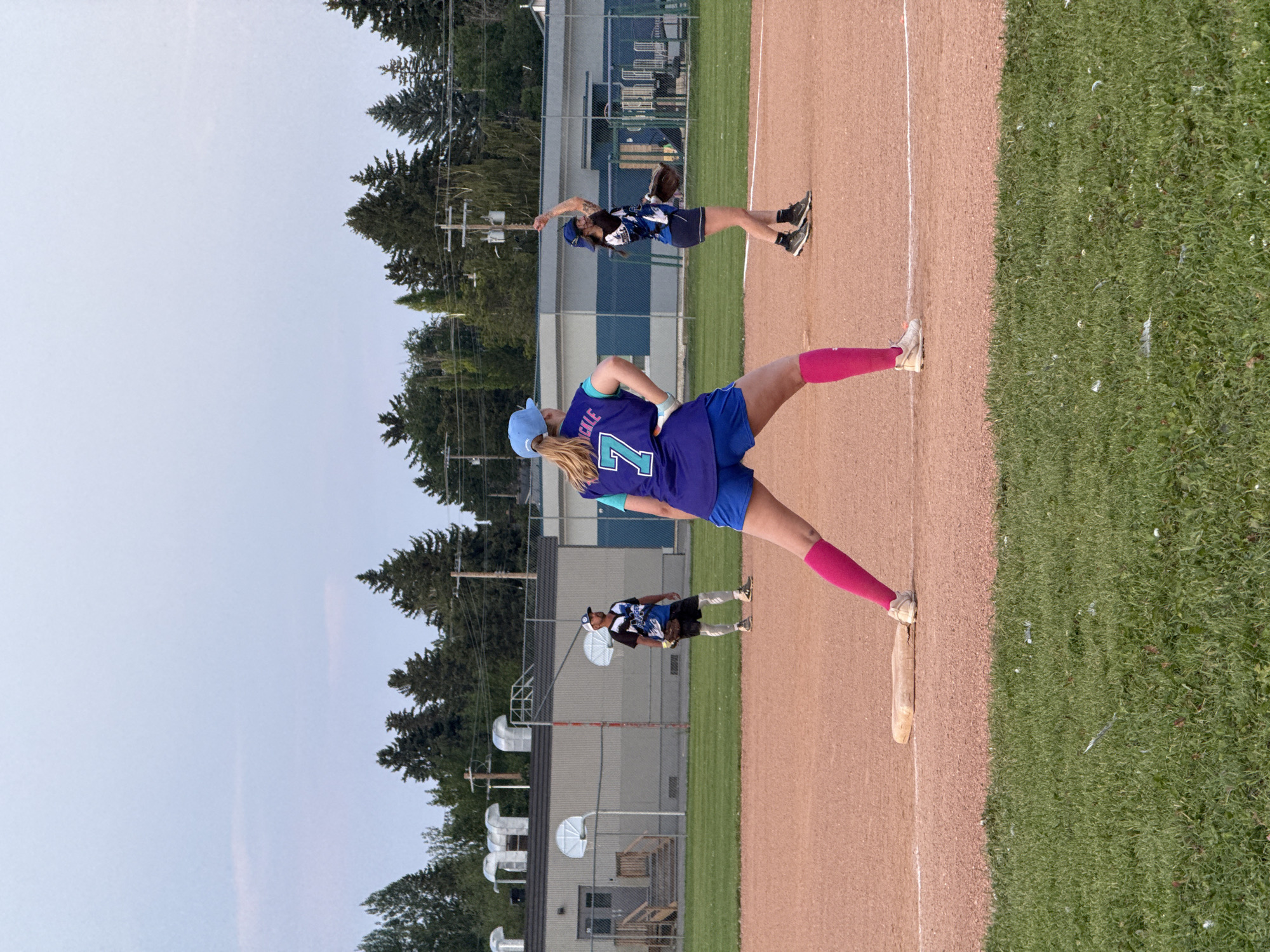 The image depicts a softball game in progress on a dirt field, likely in Calgary, Canada, given the presence of coniferous trees common to the region and the architectural style of the nearby building. The sky is overcast and light blue, suggesting it is either early morning or late evening.
In the foreground, a female softball player wearing a blue uniform with the number "7" on the back, royal blue shorts, and bright pink knee-high socks is positioned on the pitcher's mound. Her stance suggests she is preparing to pitch or has just released the ball, with her left leg extended forward and her right arm raised. A second female player in a similar blue uniform is visible in the middle ground to the right, also in a throwing or fielding stance. A male player in a black and blue jersey, shorts, and knee-high socks is in the background on the left, appearing to be fielding.
Behind the players, a light-colored building with large metal ventilation pipes is visible, possibly a school or community center. A chain-link fence surrounds the field. The ground is a mixture of dirt and grass, with a white baseline marked on the dirt. The overall scene captures a moment of athletic activity in an outdoor sports setting.