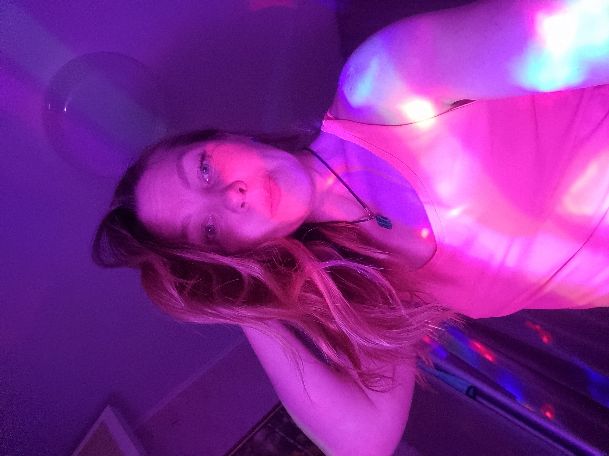 A woman is pictured indoors, likely in Chaikovsky, Russia, judging by the request context. She is illuminated by pink, purple, and blue lights, which may be party lights. The woman is wearing a pink shirt and a necklace with a pendant. Her hair is a reddish-blonde color. In the background, there is a balloon, a picture frame on the floor, and what appears to be a shiny material with colored light reflecting off it.