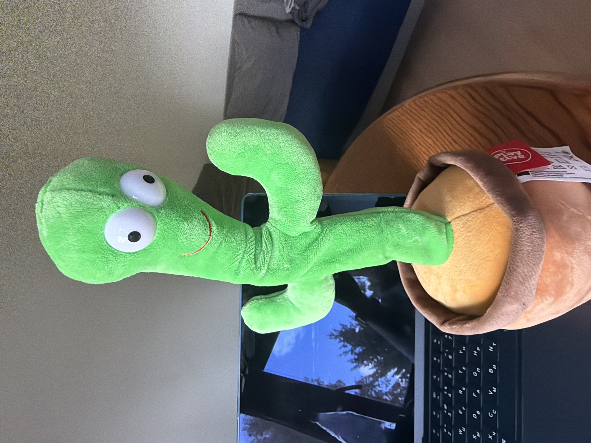 A plush toy cactus stands upright in a terracotta-colored pot on a wooden table. The cactus is bright green with large white and black eyes and a small red smile. Its arms are in a gesture resembling waving. A laptop is positioned behind the cactus, its screen displaying a reflection of trees and the sky. The background shows a couch with dark blue and grey upholstery, and a wall. The image was taken in a room in Piastów, Poland.