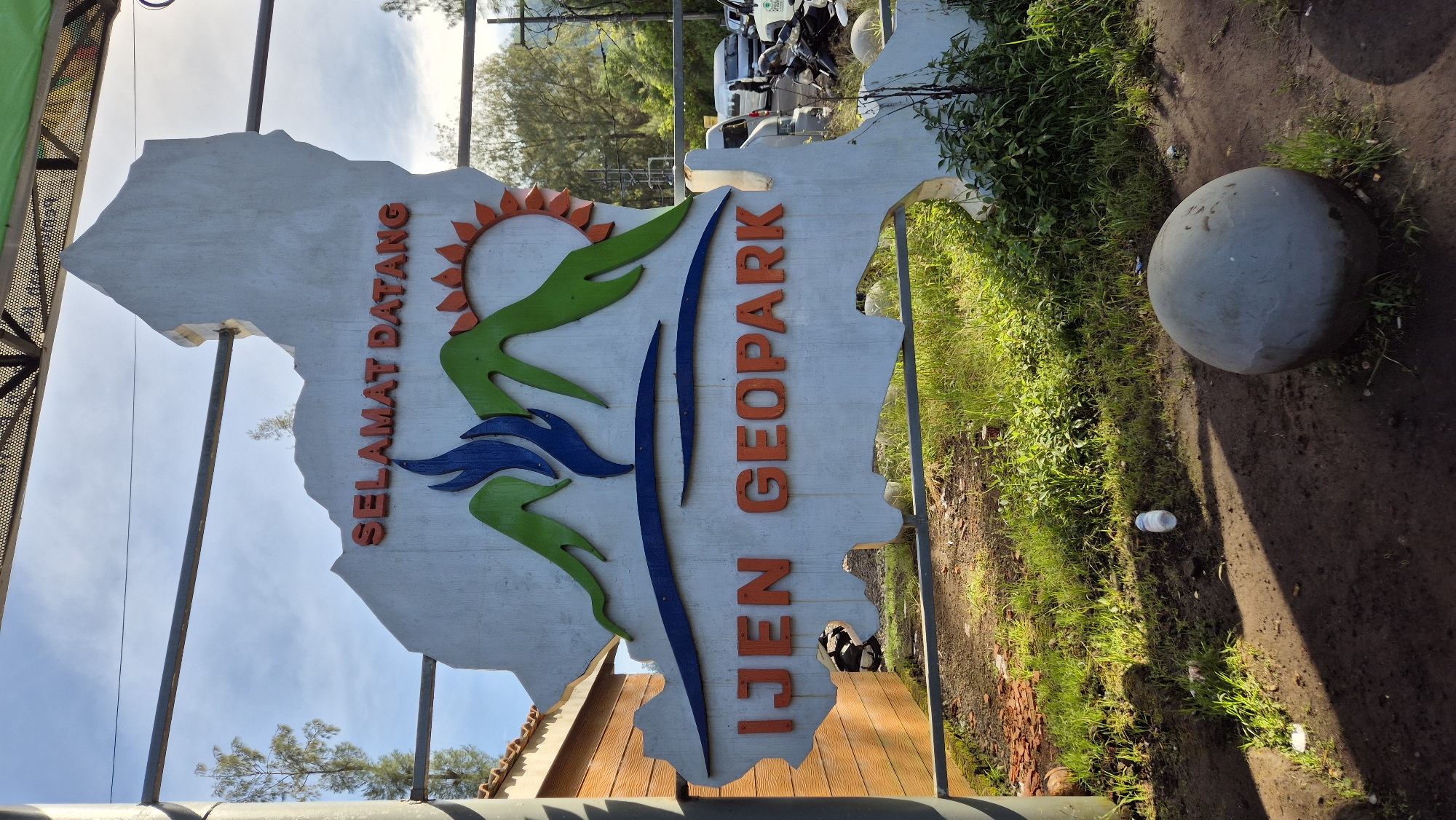 The image depicts an "IJEN GEOPARK" sign in Unknown, Indonesia. The sign is shaped like a map, bearing the words "SELAMAT DATANG" and graphic elements of a sun, green mountains, and blue waves. The sign is mounted on metal posts and is positioned next to a grassy area. To the right of the sign, a large grey concrete sphere rests on the ground. In the background, a road with parked vehicles and trees can be seen.
