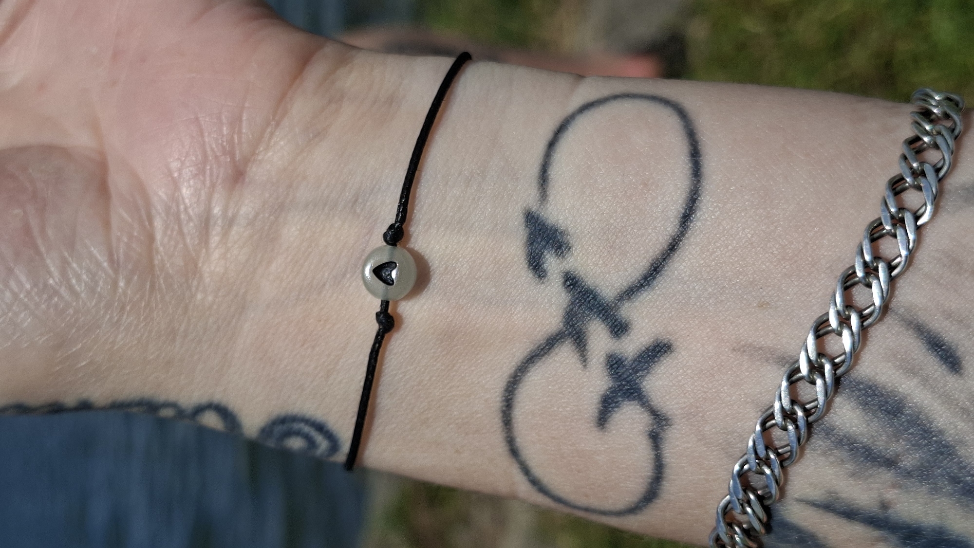 A close-up shot focuses on a person's forearm and wrist, adorned with multiple tattoos and jewelry. The skin is fair and shows wrinkles. A black string bracelet with a single, light gray, round bead is visible around the wrist. The bead has a triangular cutout. Below the bracelet, a large tattoo of an infinity symbol is inked onto the skin, with what appear to be two arrowheads integrated into its design. Further down the forearm, a silver chain bracelet is worn. The background is blurred, showing hints of water on the left and greenery on the right, suggesting an outdoor setting. The image does not provide any information about the city of Смалявічы, Belarus.