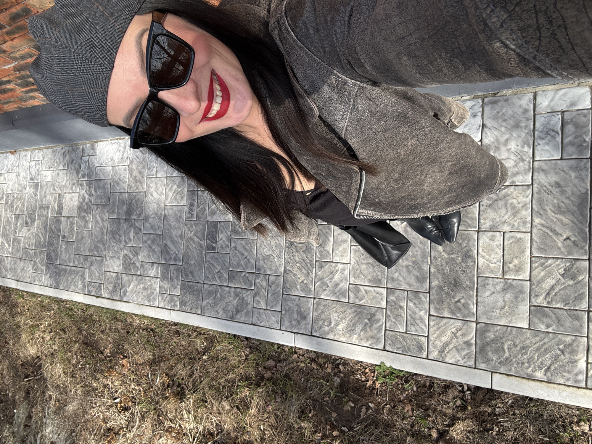 A woman in sunglasses and a patterned hat is seen from above, smiling and looking upward. She is wearing a dark jacket and black boots. She stands on a paved walkway made of gray rectangular stones. The edge of the walkway is lined with a gray border, and adjacent to the walkway is an area of brown grass and dead leaves.
I don't have information about this image or location to be Chaikovsky, Russia.