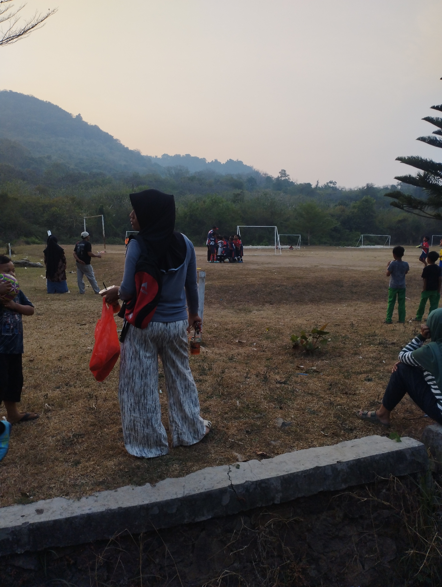 In the city of Kramatwatu, Indonesia, a group of people are gathered on a dusty, dry field with a mountainous, tree-covered landscape in the background under a pale sky. In the foreground, a woman wearing a black hijab and a grey long-sleeved shirt with patterned wide-leg pants stands facing away from the camera. She is carrying a red plastic bag in her left hand and a bottle with a straw in her right. A red backpack is slung over her shoulders. To her left, a young boy in a dark t-shirt and dark shorts holds a ball. Further back, several other individuals are visible, some near a soccer goal. Two children in green pants stand in the middle ground. Another person in a green top and dark pants is seated on a stone wall in the lower right corner of the image. The overall impression is of an outdoor gathering, possibly for a sporting event or recreational activity.