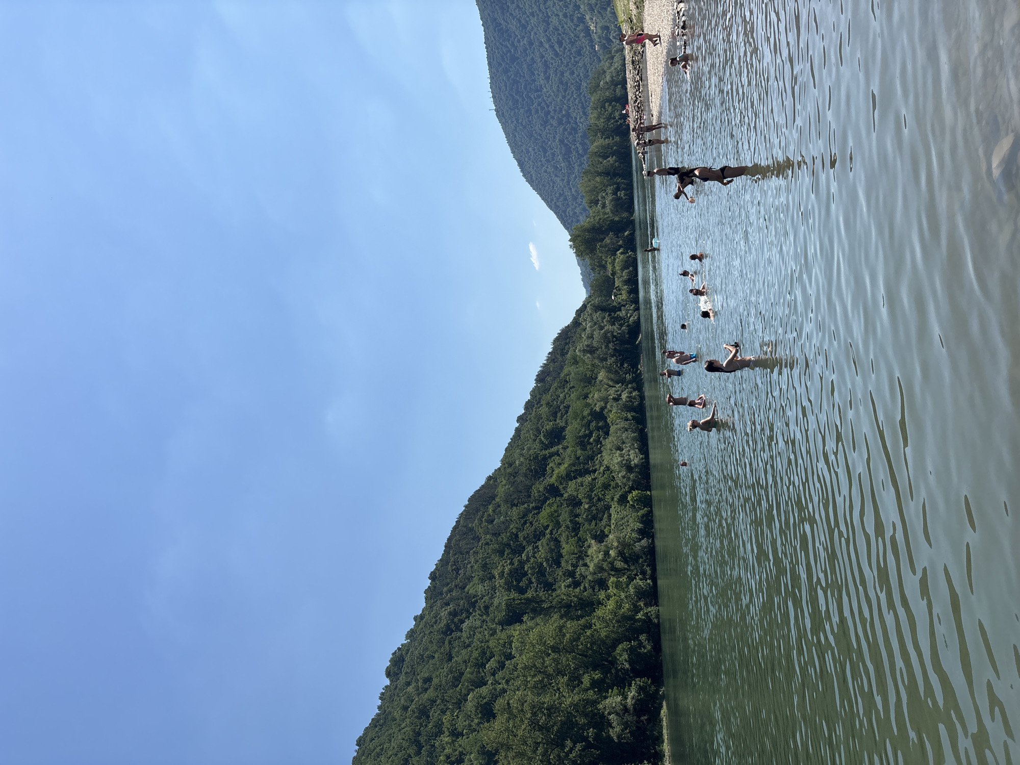 The image shows a sunny day at a body of water, likely a river or lake, with lush green hillsides rising from its banks. The water is clear and calm, with ripples on the surface. Several people are in the water, some swimming, some standing, and some appearing to be playing or relaxing. The far bank is lined with trees, and a rocky area is visible in the upper part of the image, where more people are gathered. The sky is a clear, bright blue, indicating good weather. The overall scene evokes a sense of leisure and enjoyment on a warm summer day.