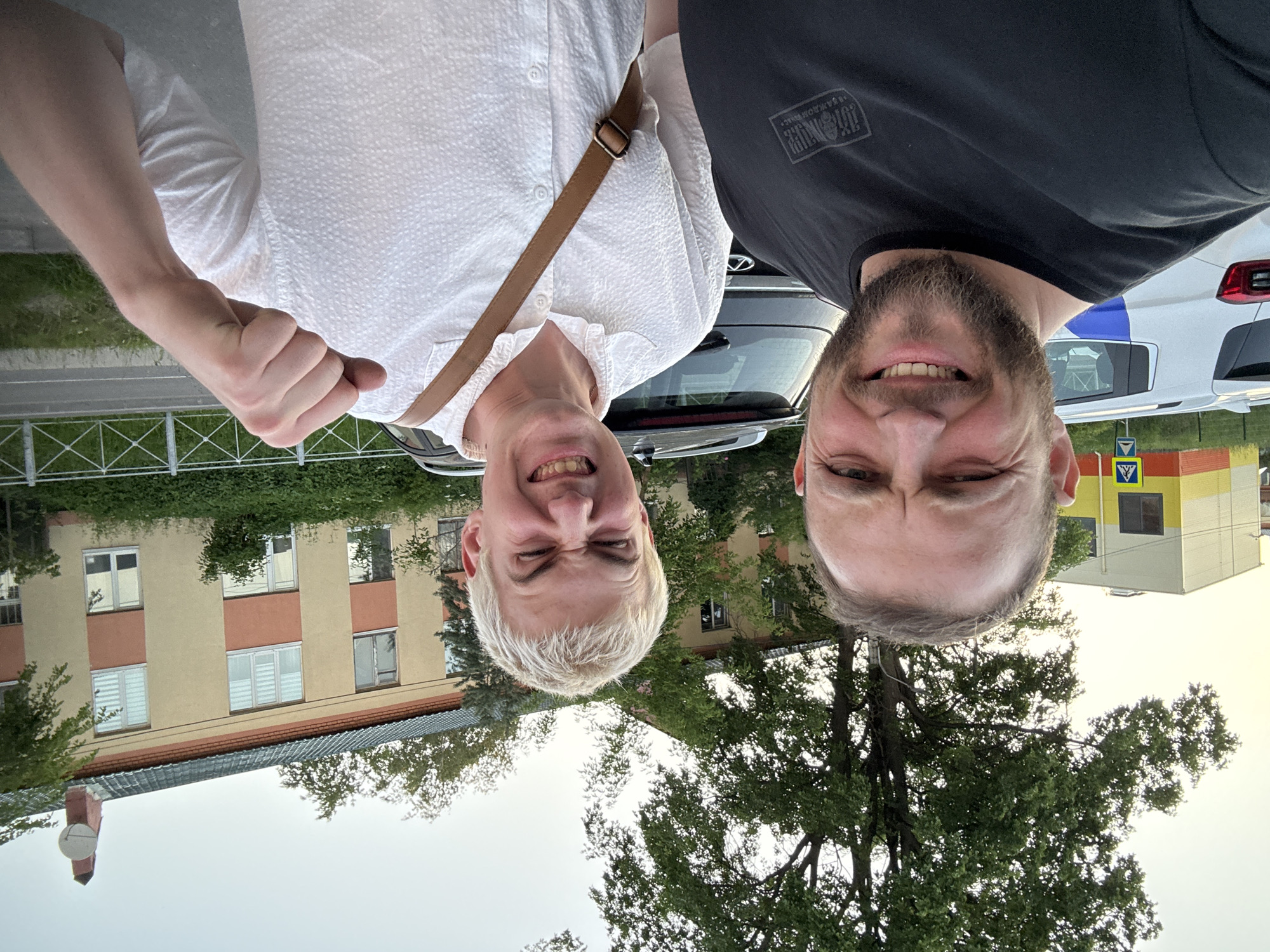 Two adult males are captured in a selfie-style photograph taken outdoors in the city of Unknown, Unknown. The lighting suggests late afternoon or early evening under a clear or partly cloudy sky.
The man on the left, closer to the camera, is a male with a short dark beard and mustache, short dark hair, and visible wrinkles around his eyes. He is smiling broadly, looking directly at the camera, with his left eye appearing slightly squinted. He wears a black short-sleeved t-shirt with a rectangular graphic design on the chest, which includes what appears to be Cyrillic script.
The man on the right, positioned slightly behind and to the right of the first man, is a male with very short, bleached blonde hair and a pale complexion. He is making an exaggerated facial expression with squinted eyes, a scrunched nose, and an open mouth revealing teeth. His right arm is bent at the elbow, with his hand clenched into a fist and held near his shoulder. He is wearing a white short-sleeved collared shirt with a textured, possibly dimpled or seersucker, fabric. A brown leather strap, likely from a bag, crosses his chest from his right shoulder to his left hip.
The background features several structures and natural elements. To the far left, a modern, multi-story building with a light yellow upper section and a red-orange lower stripe is partially visible. In front of this building, a blue pedestrian crossing sign with a white triangle and walking figure is present. Behind the men, a white SUV with blue markings on its side is parked to the left, and a dark-colored SUV is parked to the right. Both vehicles have visible roof rails. Dominating the right side of the background is an older, multi-story building with a light brown/tan facade and horizontal red-orange accent stripes. This building has multiple windows, some fitted with white blinds. Its roof is dark green, featuring a reddish chimney on the far right topped with a white satellite dish. Large, leafy green trees are visible throughout the background, particularly behind the buildings and overhanging the scene. A paved road is visible to the far right, separated from a grassy area by a white metal fence or railing.