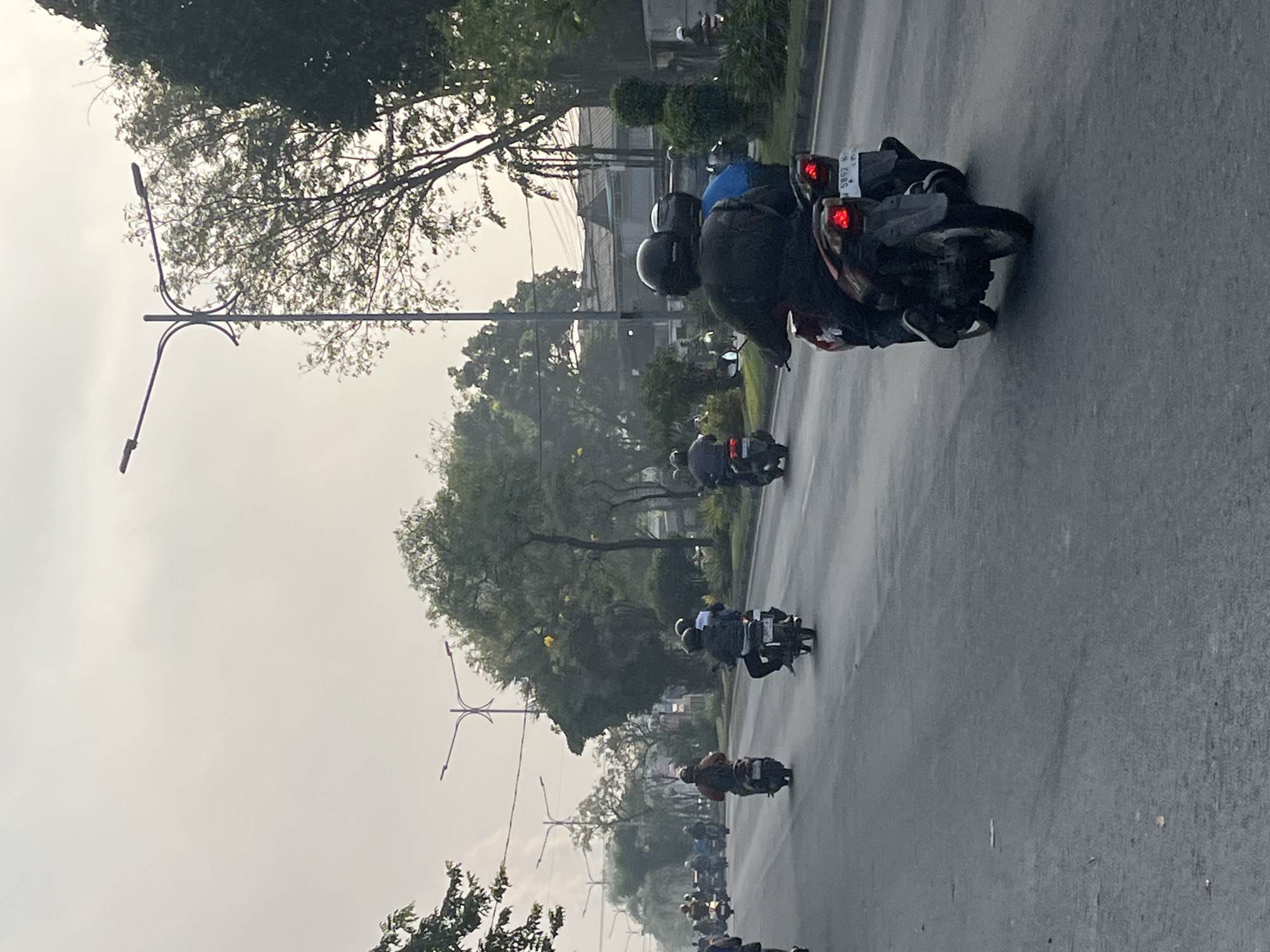 The image depicts a street scene in Gelam, Indonesia, likely during the daytime with overcast skies. A multi-lane asphalt road is visible, with several motorcycles and their riders traveling in the same direction. The motorcycles are varied in type and color, and the riders are wearing helmets for safety. In the foreground, a rider on a red and black motorcycle is clearly visible, as is a rider on a blue motorcycle behind them. Further down the road, more motorcycles can be seen, creating a sense of traffic.
The environment includes trees lining the sides of the road, some of which are dense and leafy, while others appear more sparse. Streetlights are also present, suggesting an urban or suburban setting. In the background, buildings can be seen, including some with tiled roofs, typical of Indonesian architecture. The overall impression is one of a typical day on the road in a busy area of Gelam, Indonesia, with people commuting on motorcycles.