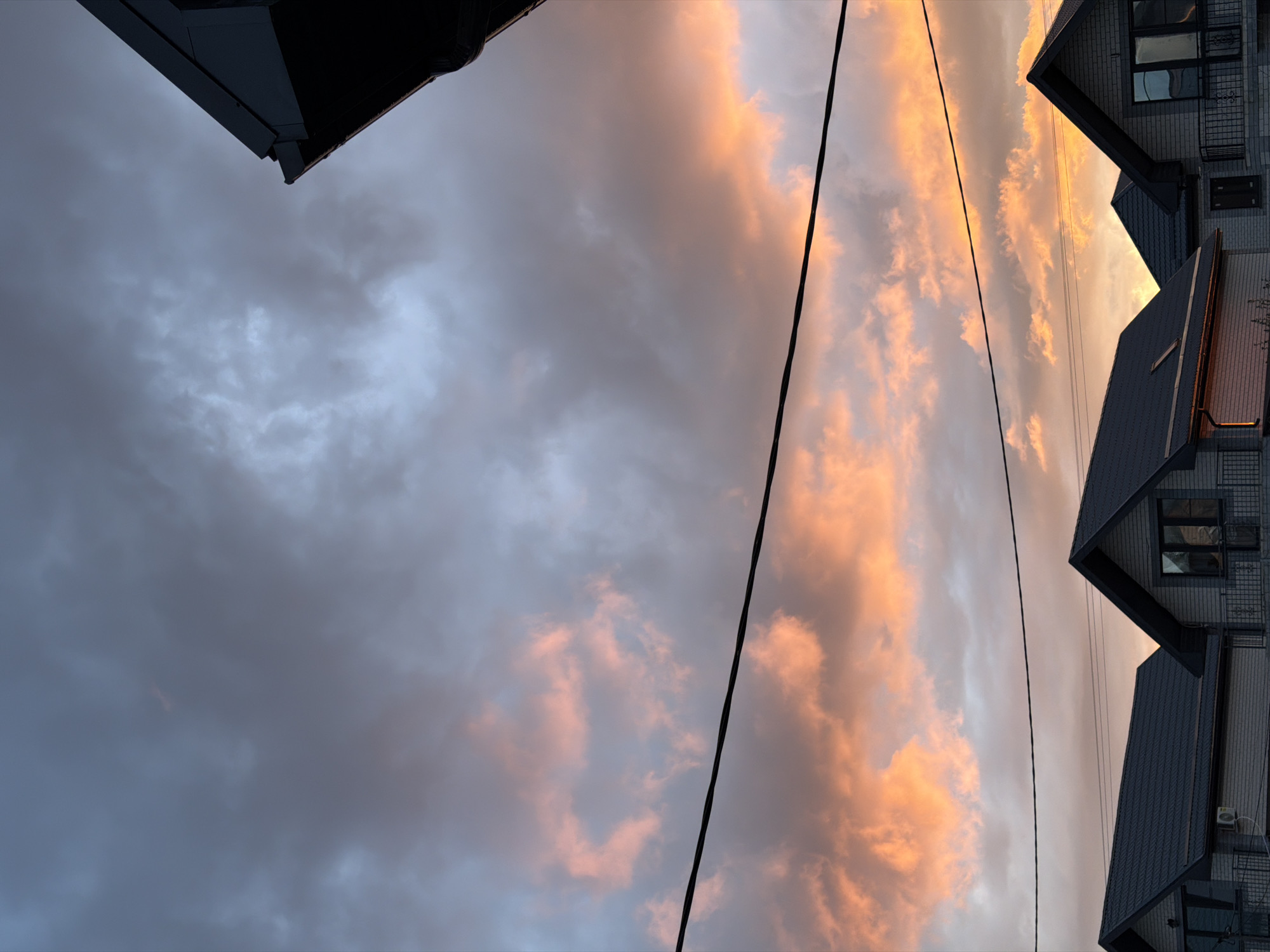 The image depicts a sky at sunset in Rossiyskiy, Russia, partially obscured by buildings and power lines. The sky transitions from a deep blue-gray to orange-pink hues as the sun sets. Power lines bisect the image diagonally, obscuring the view. A fragment of a dark roofline is visible in the upper left corner, while buildings, with their dark roofs and light walls, are seen in the upper right. Windows are also seen in the buildings.