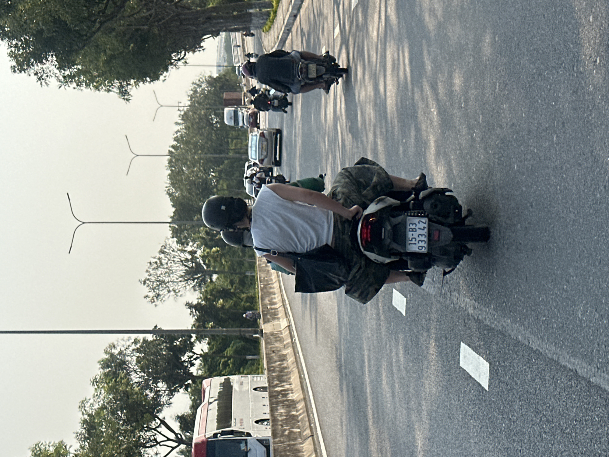 A medium shot of a two-lane highway in Đà Nẵng, Vietnam, shows two people on a motor scooter, traveling in the right lane. The driver of the scooter wears a black helmet and a white t-shirt, and the passenger wears military-style, camouflage shorts. The Vietnam license plate on the scooter reads “15-B3 933.47”. The edge of the two-lane highway is lined with a concrete barrier and foliage. A tour bus travels in the lane adjacent to the scooter. Overhead streetlights and electrical wires are also visible. In the distance, cars, other motor scooters, and a truck travel on the highway. Foliage, trees, and a hazy sky appear in the background.